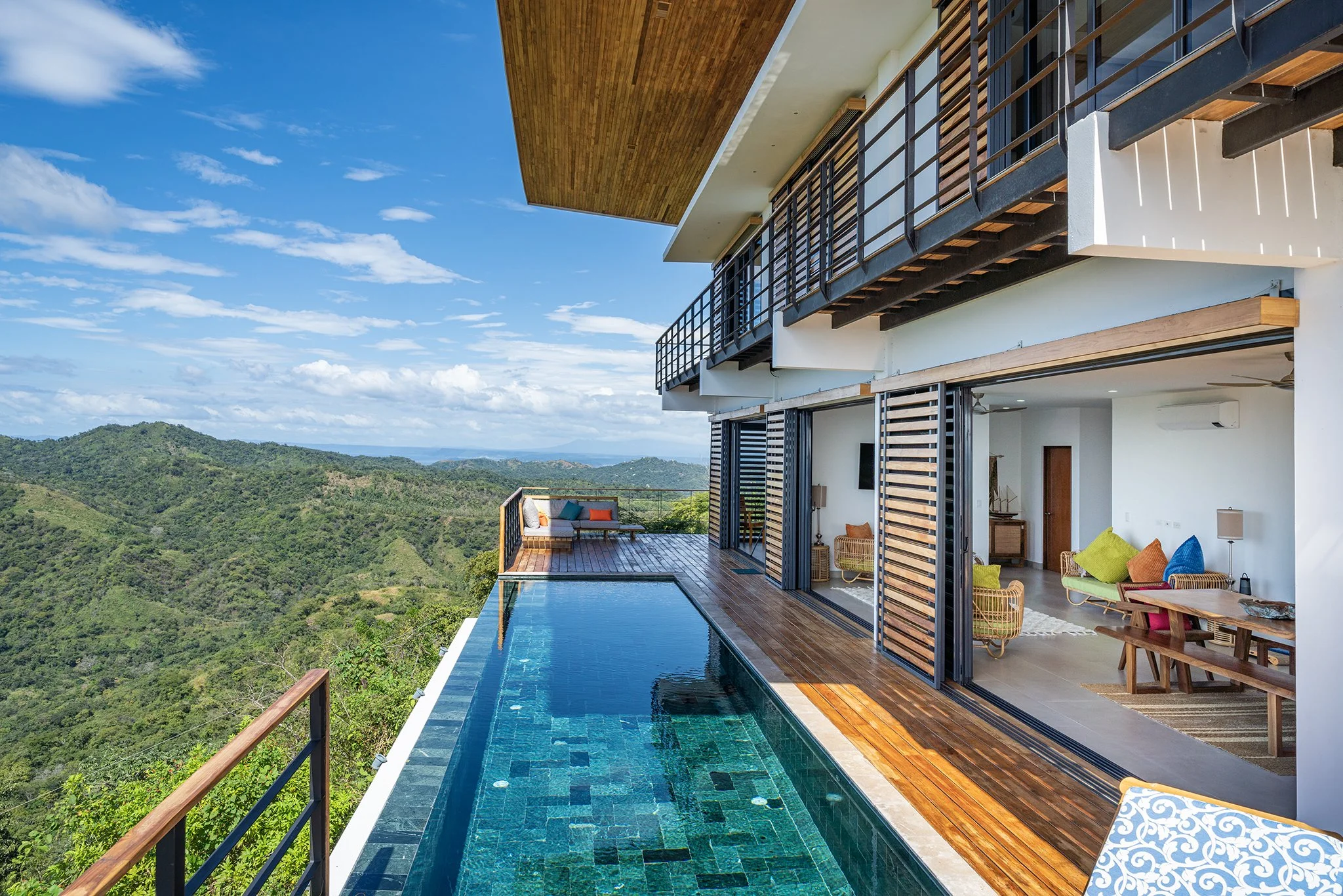 Modern house with an outdoor infinity pool overlooking green mountains under a partly cloudy sky, featuring a terrace with colorful outdoor furniture and wooden accents.