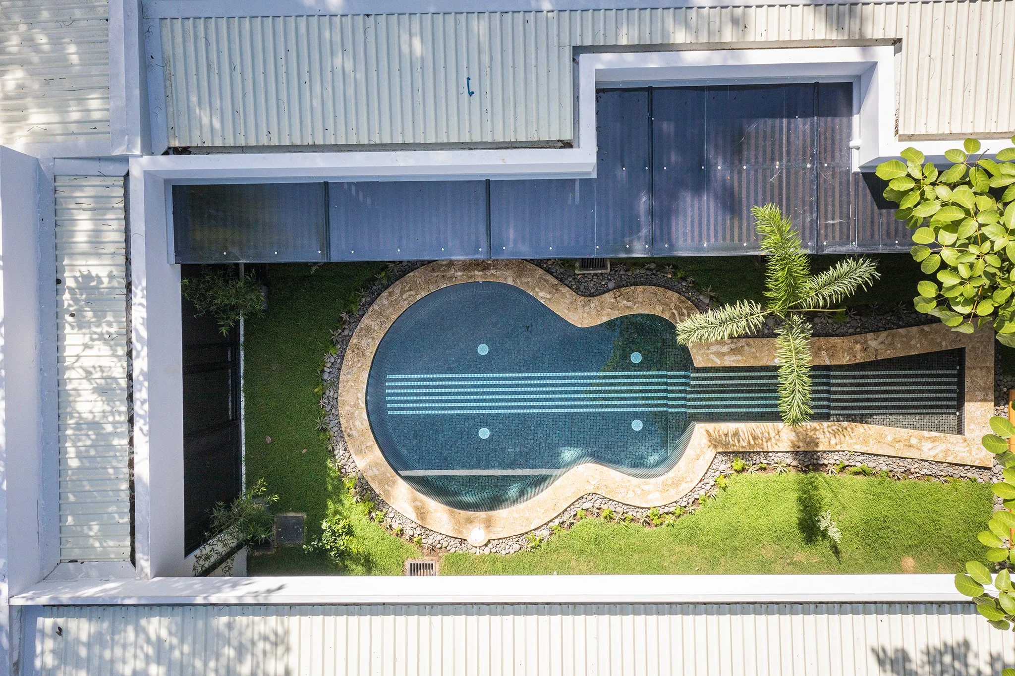 Aerial view of a backyard with a kidney-shaped swimming pool, surrounded by stone pavers, lush green grass, and tropical plants, with a modern house featuring a white siding exterior.