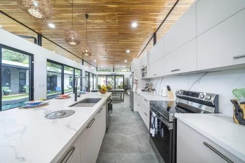 Modern kitchen with white cabinets, marble countertops, black stove, and wooden ceiling, with large windows and outdoor view.