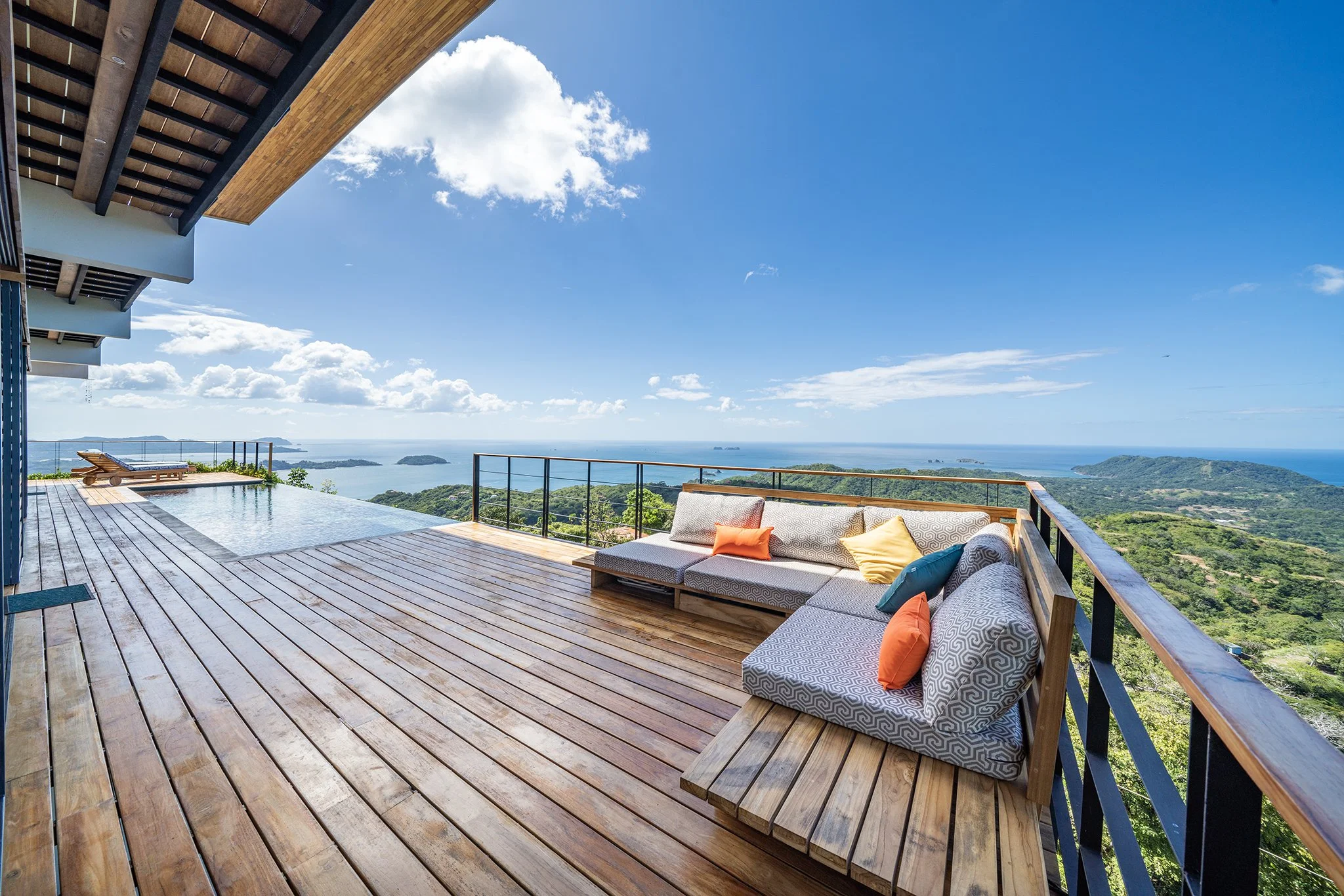 A spacious outdoor wooden deck with seating, overlooking a scenic view of mountains, the ocean, and islands under a bright blue sky with some clouds.