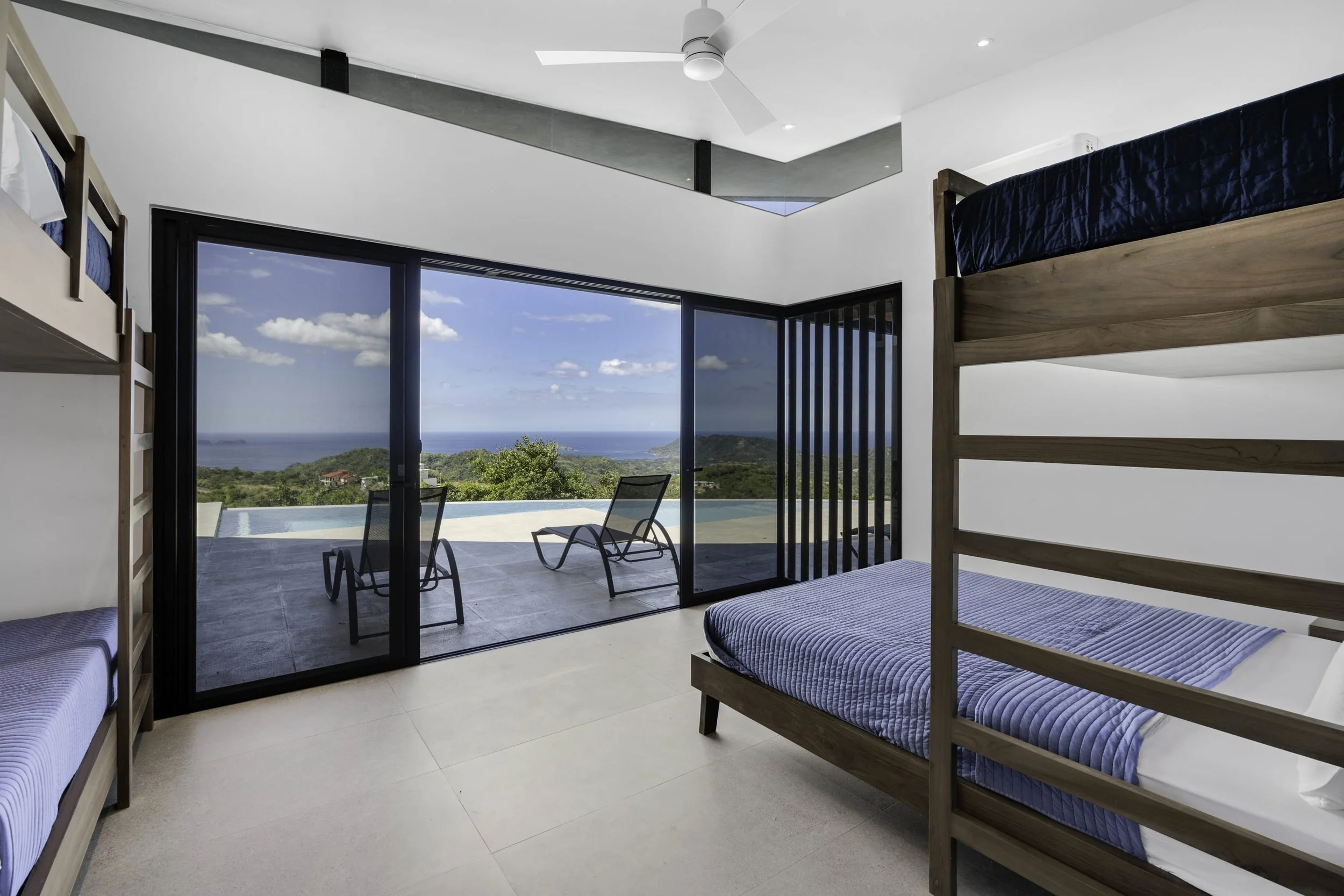 Bedroom with wooden bunk beds with blue mattresses, large sliding glass doors leading to a balcony with two chairs, and a view of the ocean and green hills.