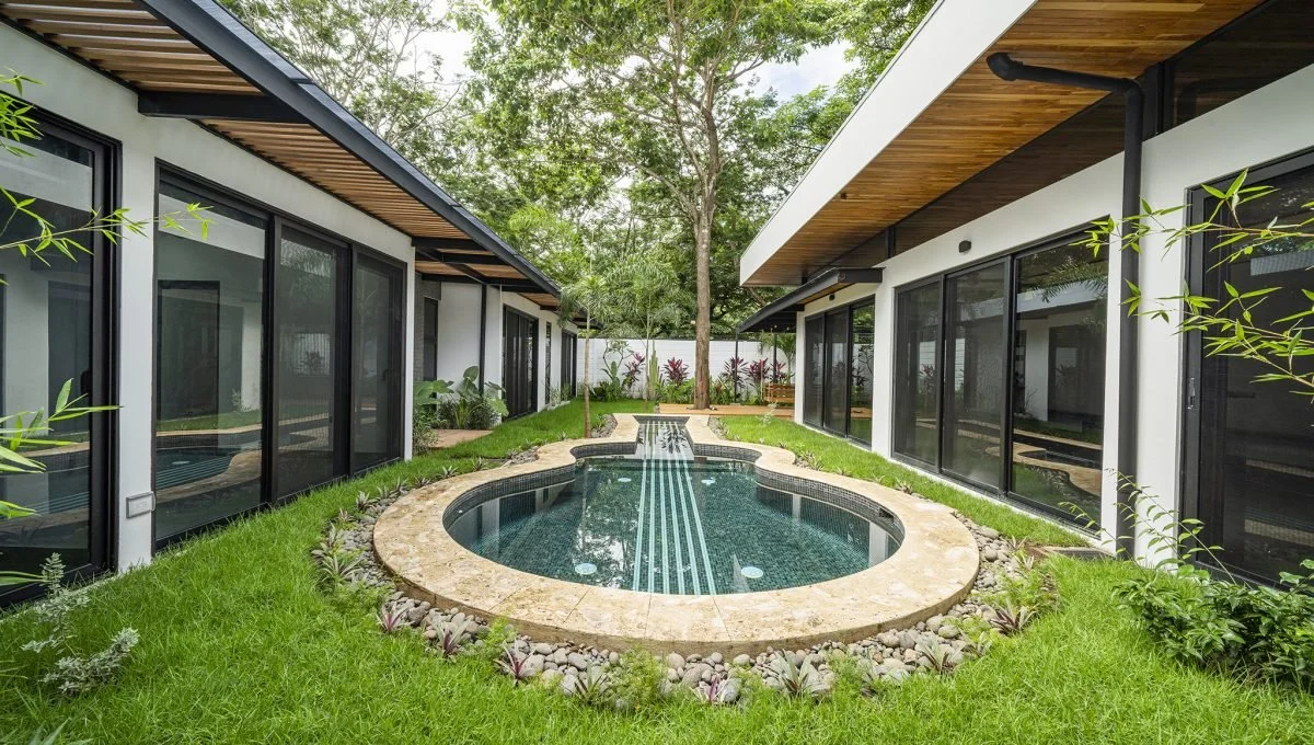 Modern backyard with a small, kidney-shaped pool surrounded by lush green grass and plants, with two contemporary houses on either side featuring large glass sliding doors and wooden overhangs, and a tree in the background.