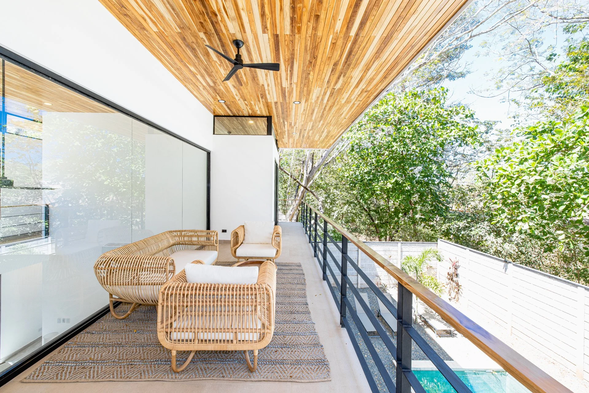 Balcony with woven rattan seating, glass sliding doors, wooden ceiling, and trees outside.