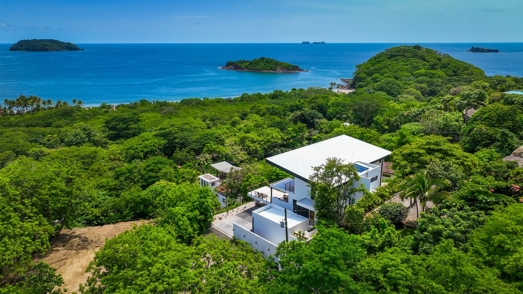 Aerial view of a tropical coastline with lush green trees, white modern house, sandy beach, and small islands in the blue ocean under a partly cloudy sky.