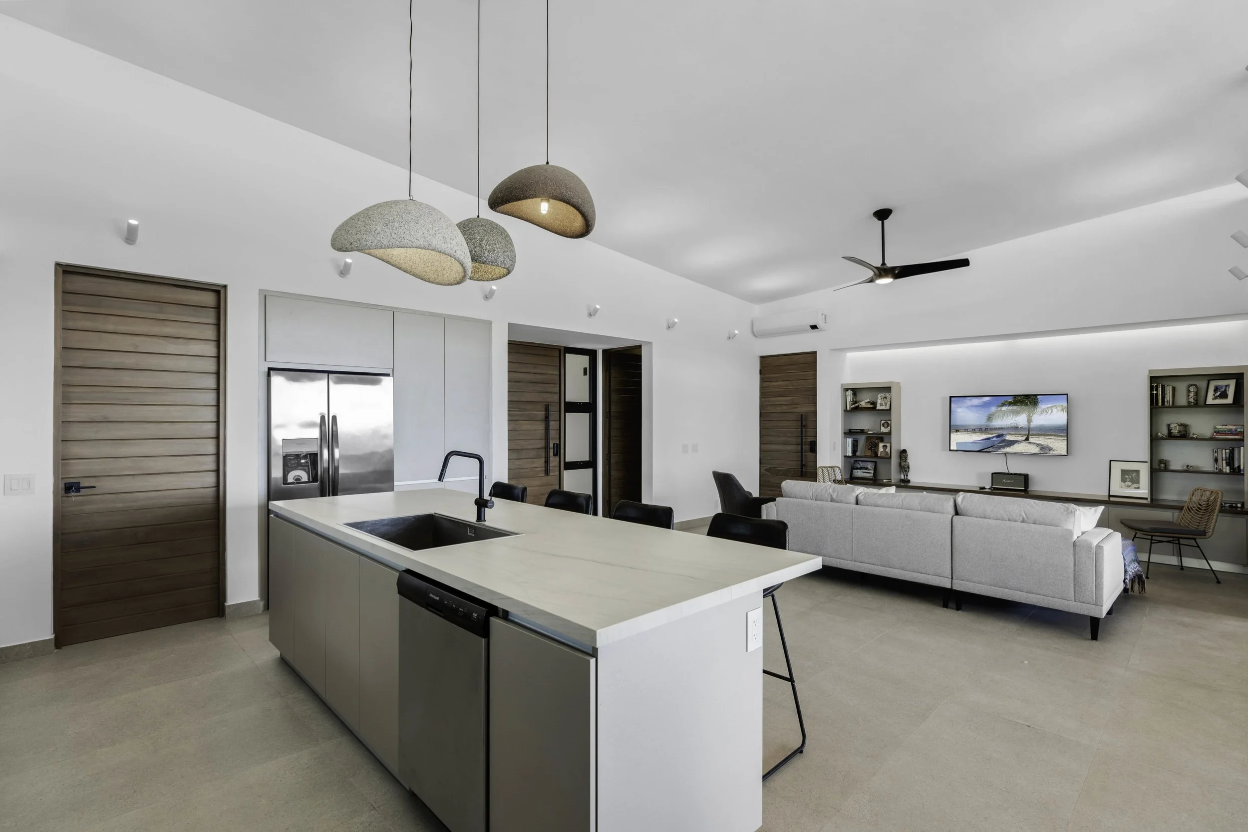 Modern open-concept living space with a kitchen island, white walls, minimalist decor, a ceiling fan, pendant lights, and a television in the background.