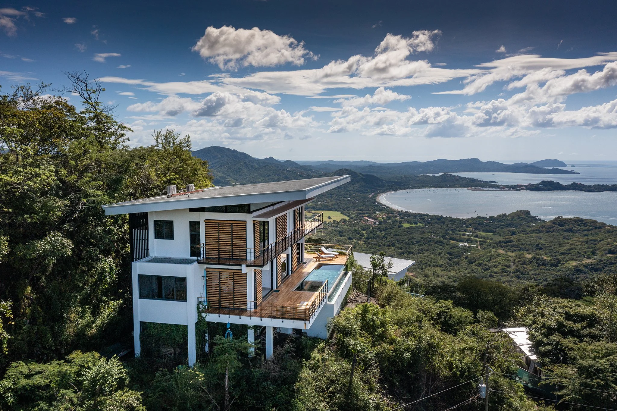 Modern house on a hillside overlooking a lush green landscape, mountains, and water with a cloudy sky.