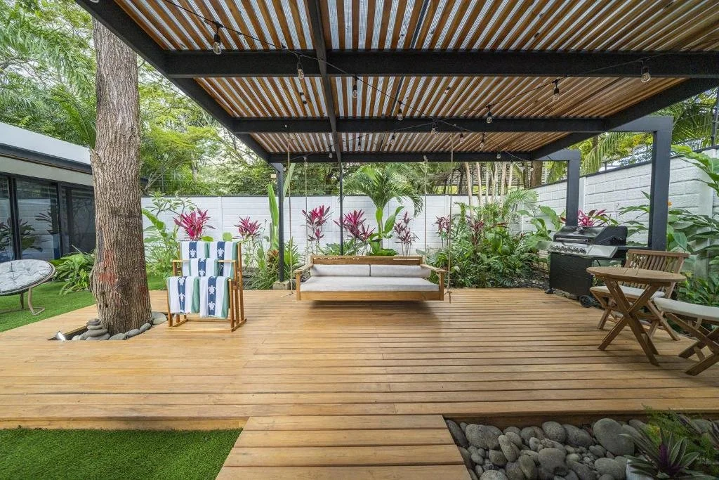 Outdoor deck area with a swing, lounge chairs, potted plants, and a barbecue grill, surrounded by a garden and trees.