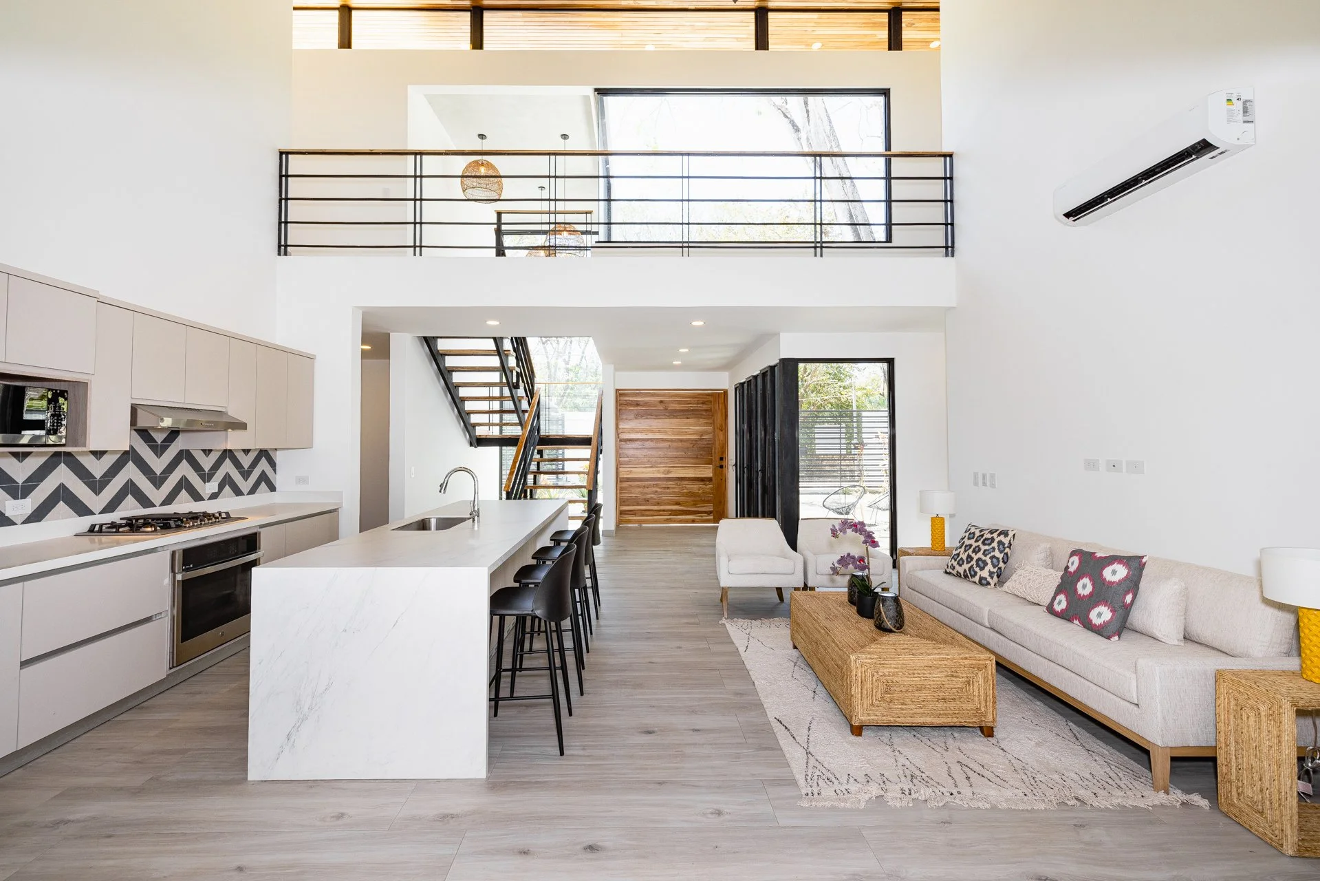 Open concept kitchen and living room with white walls, light wood flooring, modern appliances, a kitchen island with black barstools, a beige sofa with patterned throw pillows, a coffee table, and large windows letting in natural light. Staircase lea