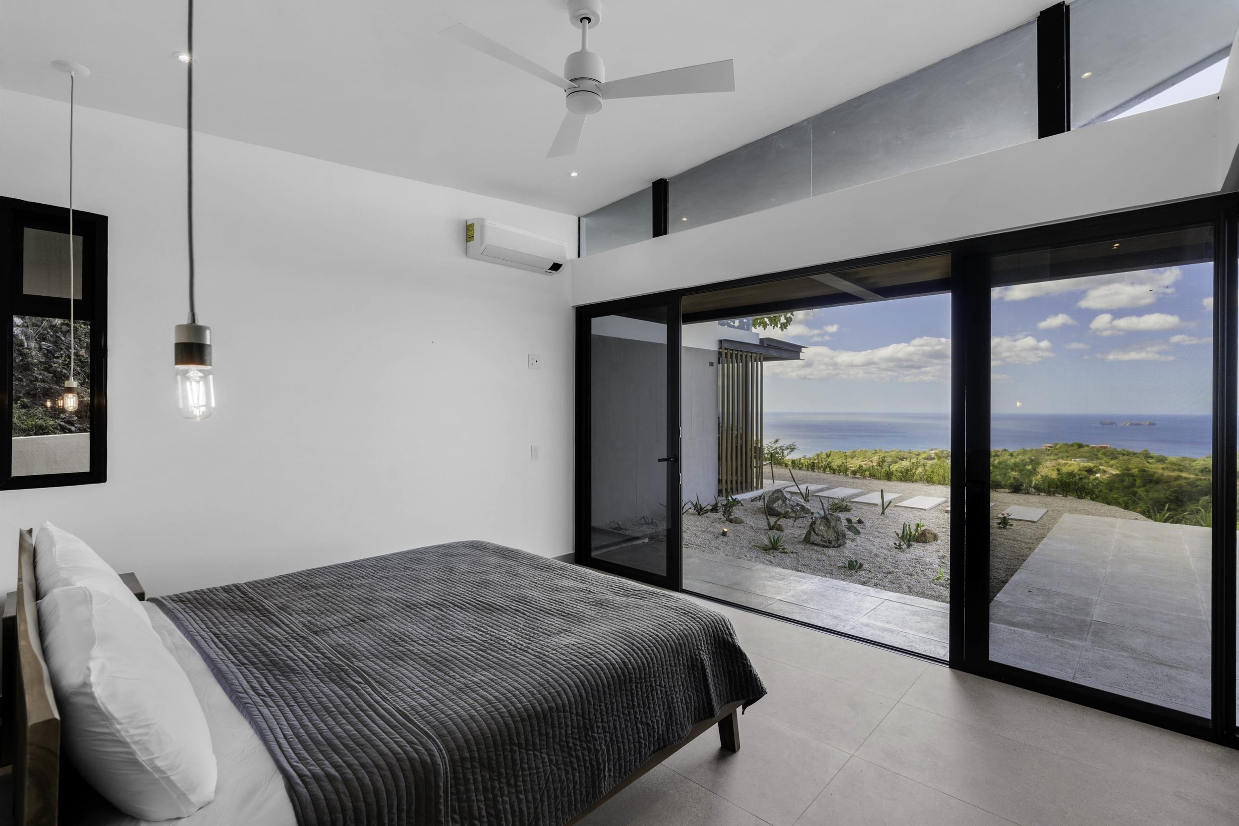 A modern bedroom with a large sliding glass door that opens to a view of the ocean and sky, a white ceiling fan, pendant lights, and a bed with a gray quilt and white pillows.