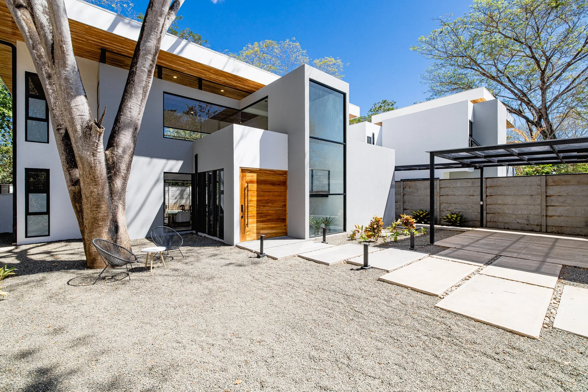 Modern white house with large glass windows, surrounded by trees and a gravel yard, featuring outdoor chairs and a concrete pathway.