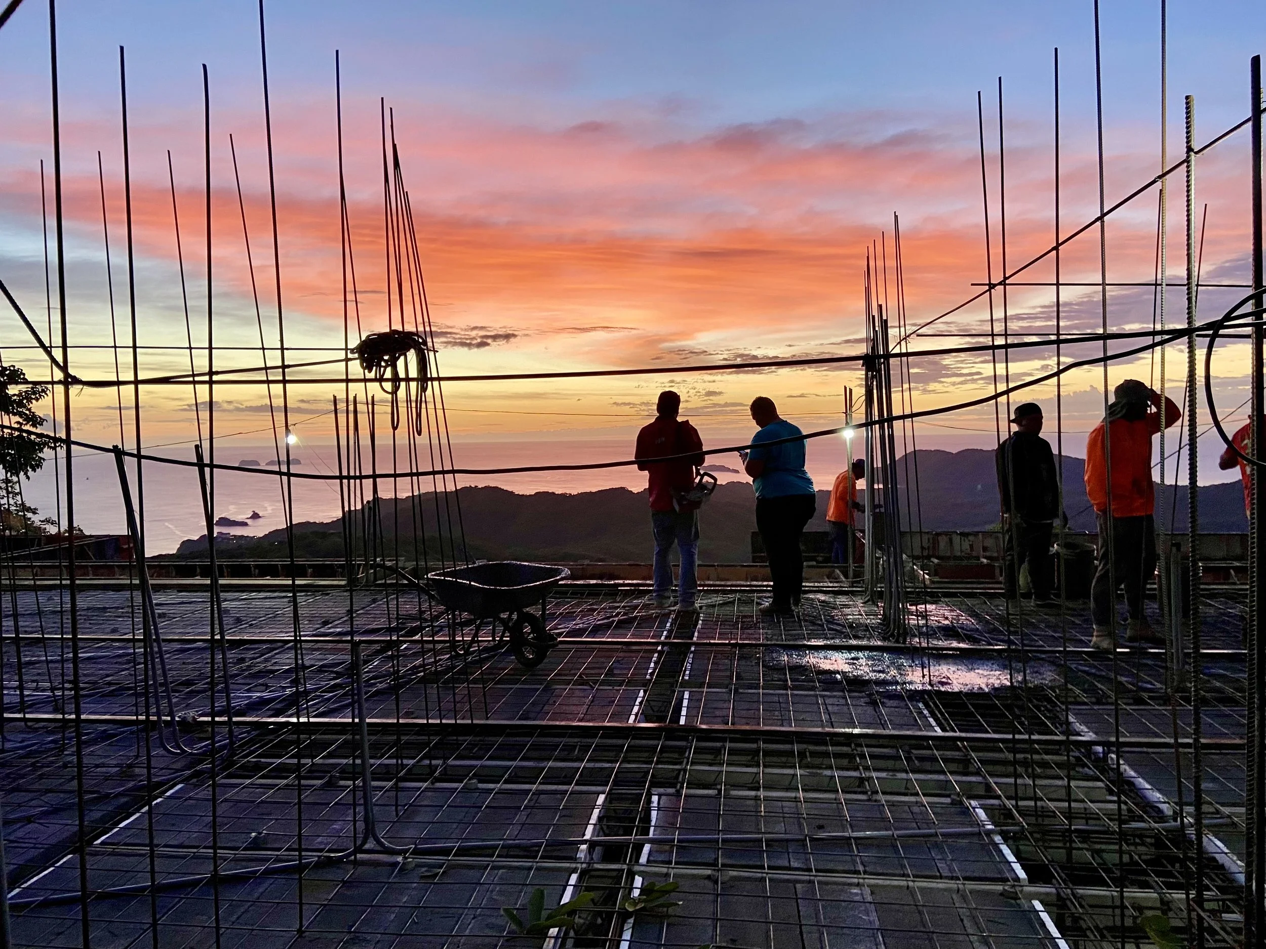 Silhouettes of people standing among metal frameworks during a colorful sunset with pink, orange, and purple clouds over water and distant land.