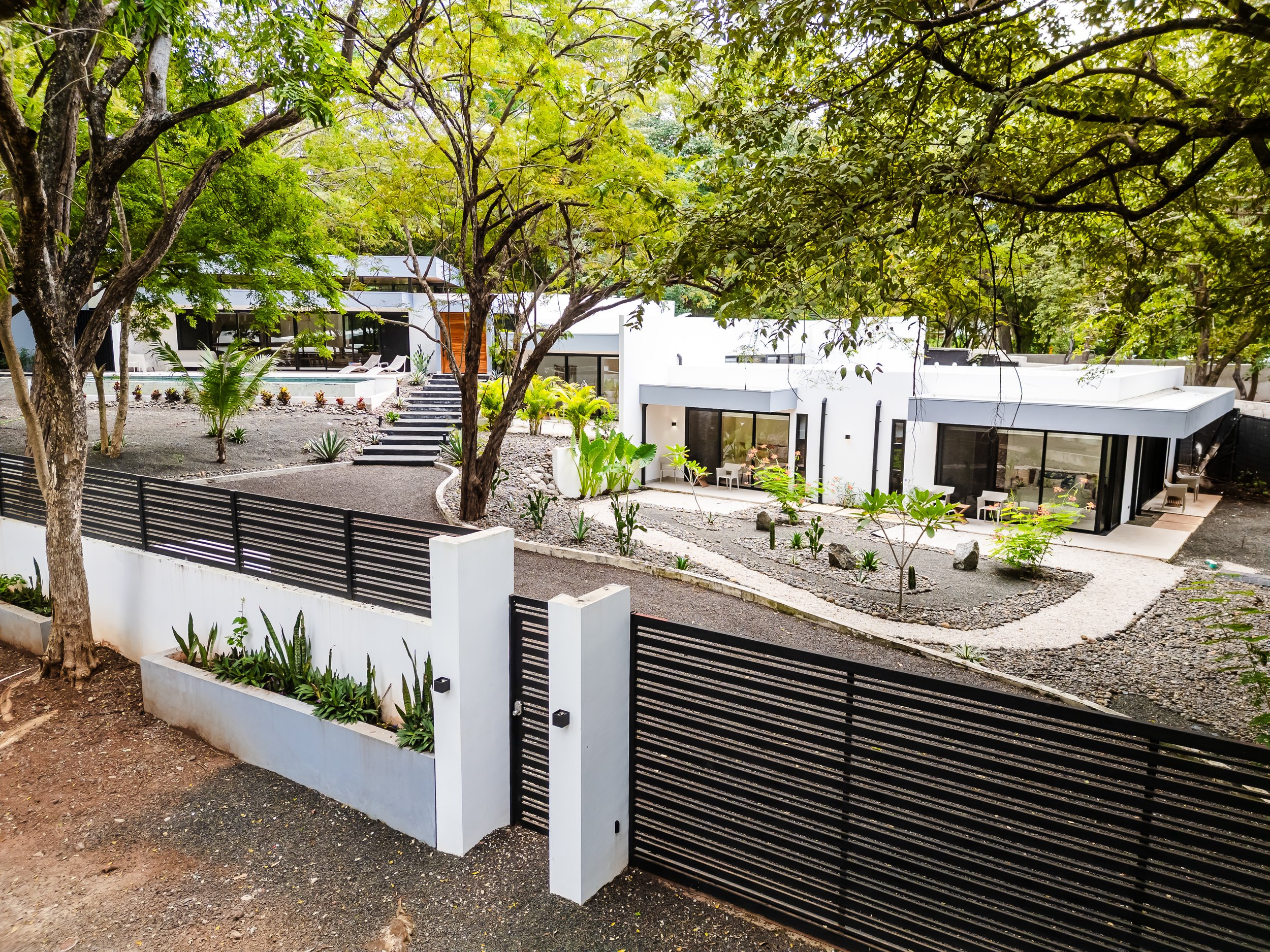 Modern white house with large windows, surrounded by trees and plants, and a black fence in the foreground.