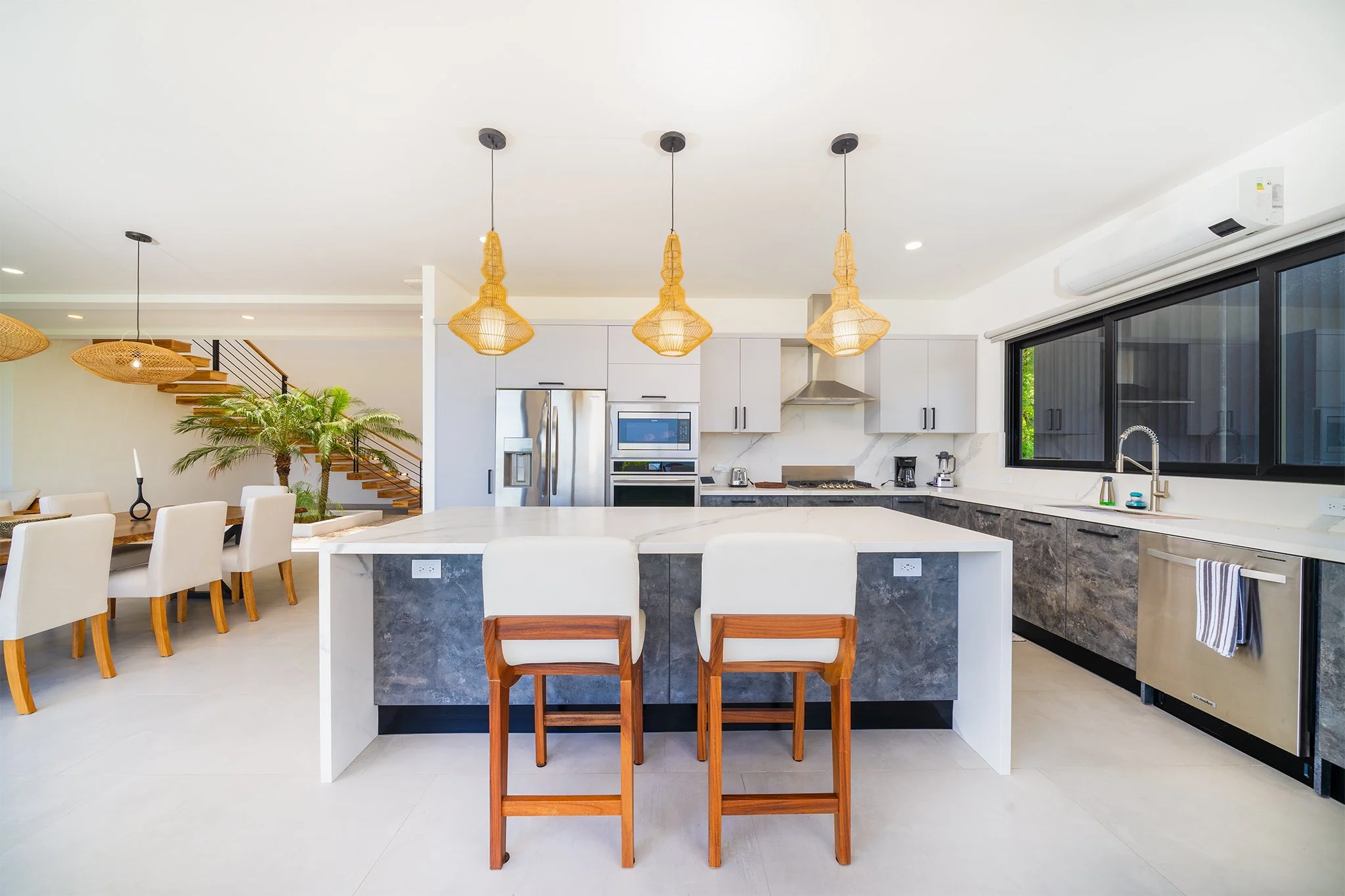Modern kitchen with white cabinets, stainless steel appliances, and a large island with seating. There are three pendant lights hanging above the island, large window with a view outside, and a staircase with wooden steps and metal railing.