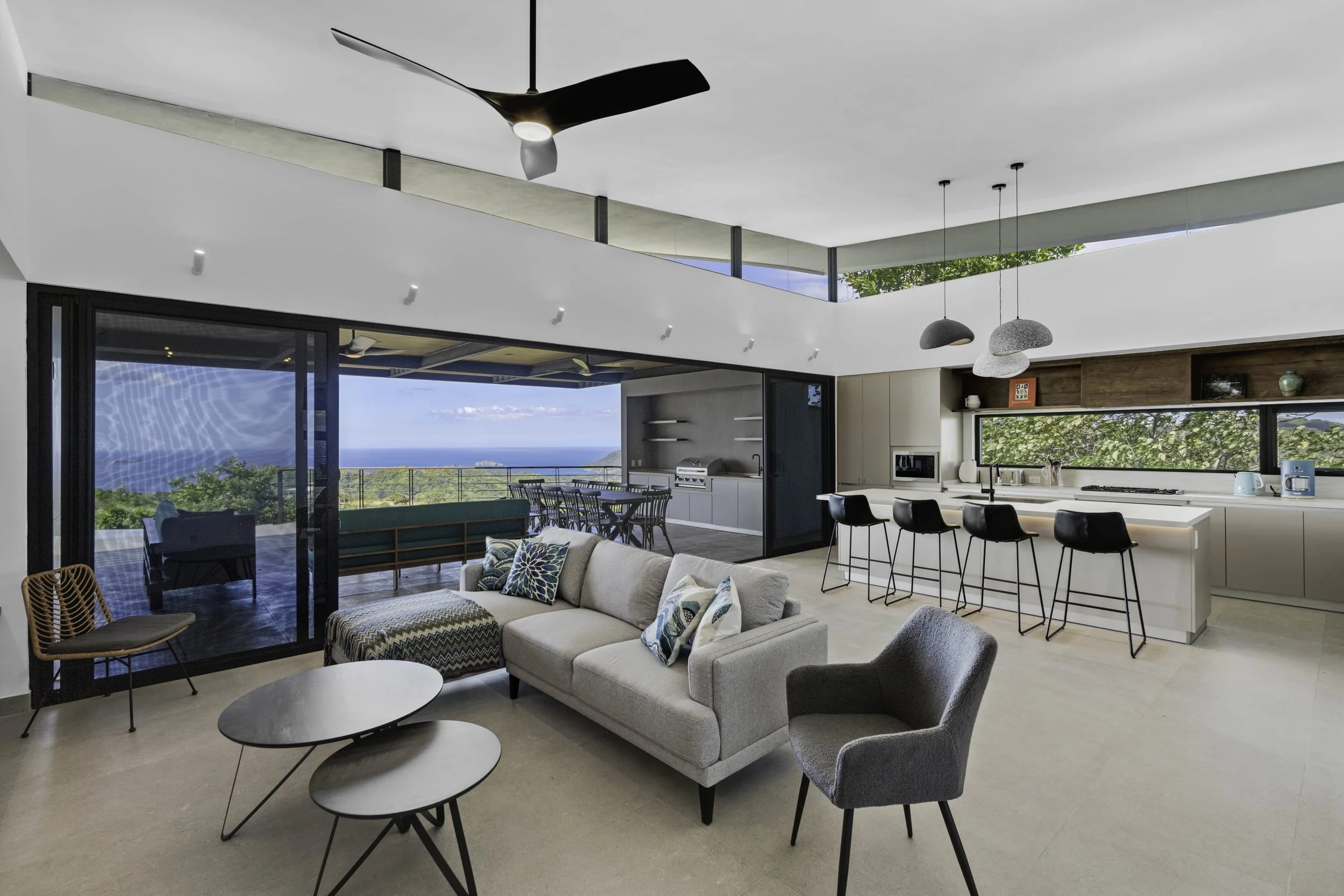 Open-concept modern living room and kitchen with large glass doors showing a view of trees and the ocean, featuring neutral-colored furniture, pendant lights, and indoor plants.