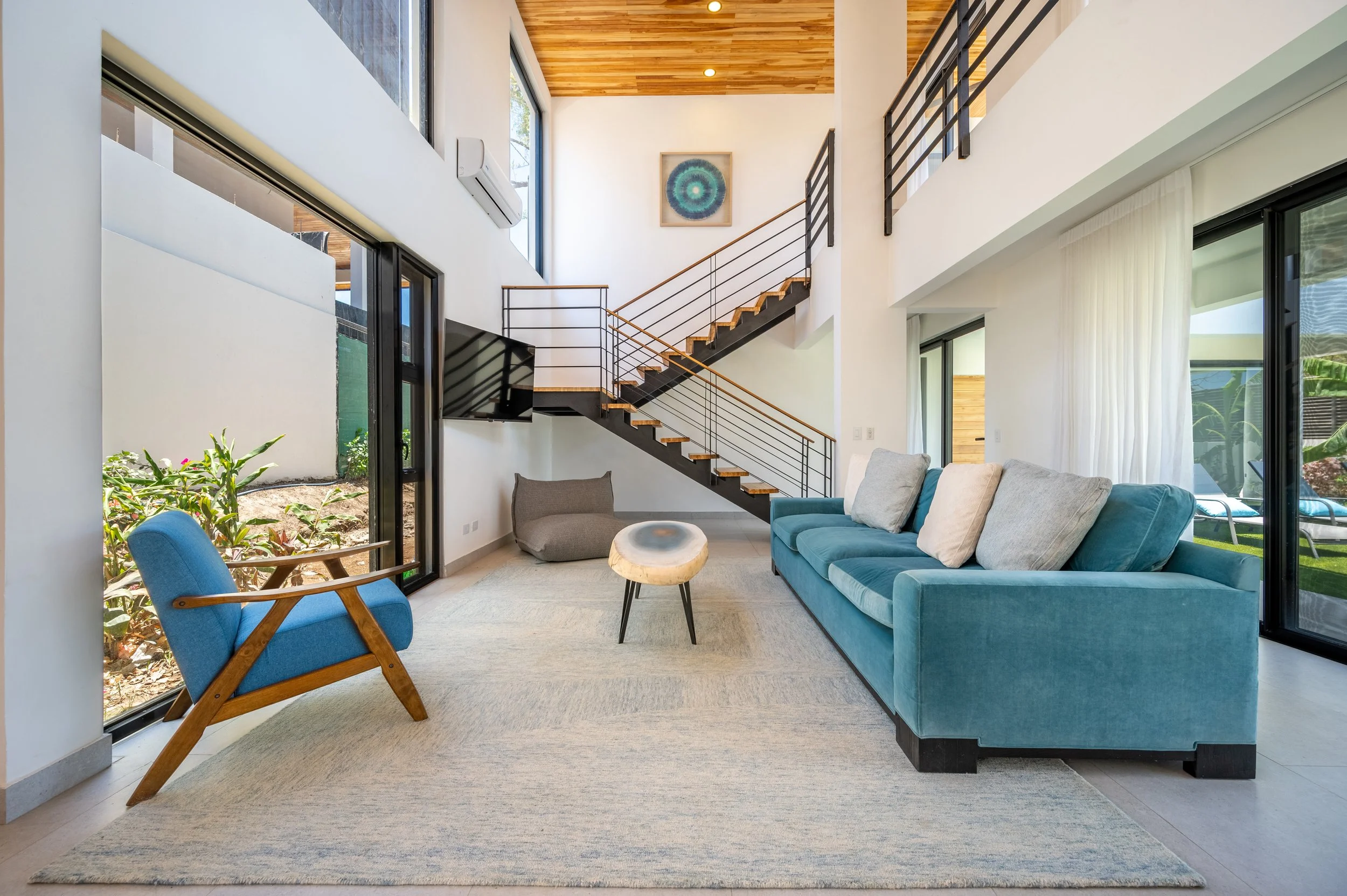 Modern living room with large windows, blue and beige sofas, a round wooden coffee table, a blue armchair, and a staircase with black risers and wooden steps leading to an upper level. Wall art and curtain accents are visible.