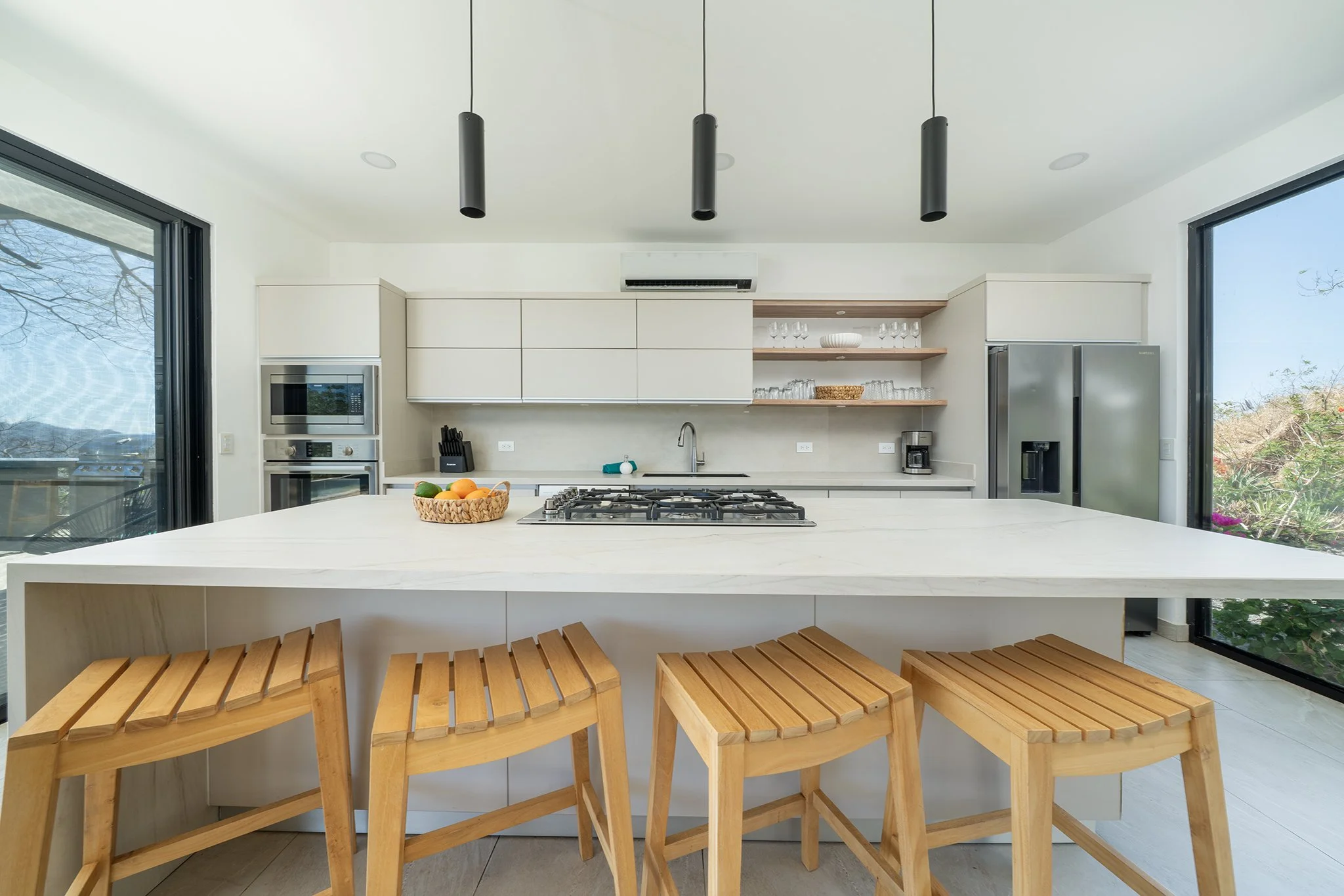 Modern kitchen with white cabinetry, open wooden shelves, and large sliding glass doors showing an outdoor view. There is a white island with a bowl of fruit, wooden stools, and a basket of fruit on the counter.