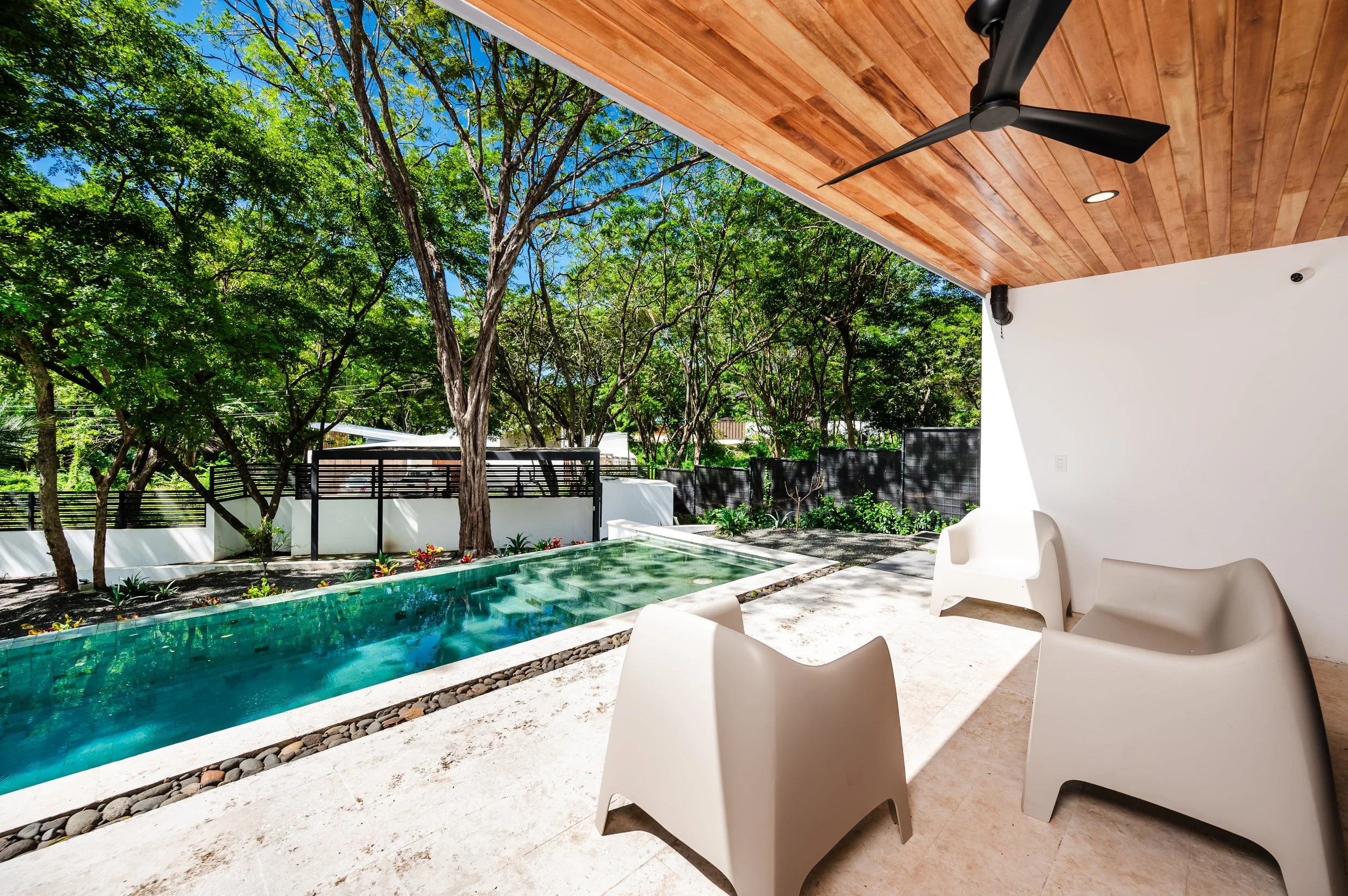 Modern outdoor patio with white chairs overlooking a swimming pool surrounded by rocks and lush green trees under a clear blue sky, featuring a wooden ceiling fan and part of a deck with warm wood flooring.