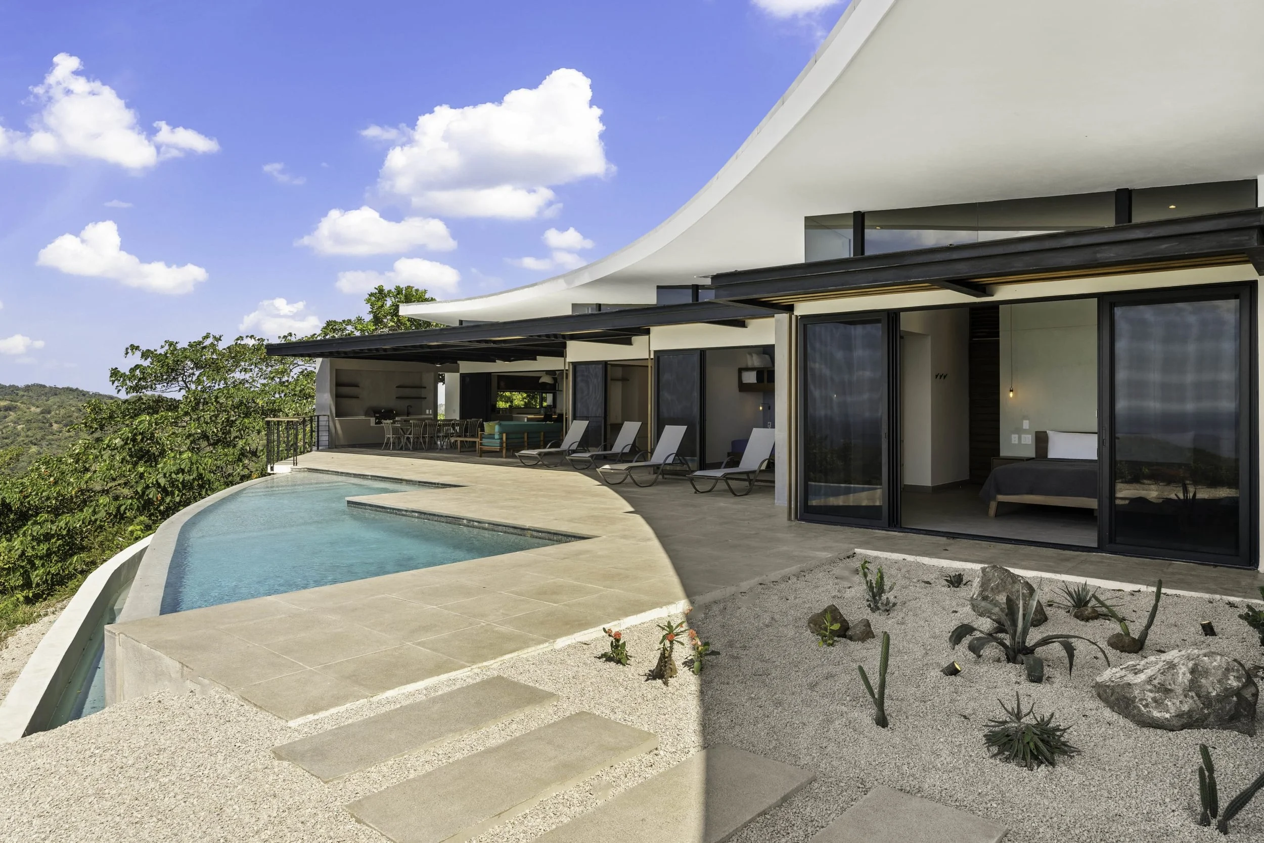 Modern white house with black accents, a swimming pool, and a patio with outdoor furniture, overlooking a green landscape and blue sky with clouds.
