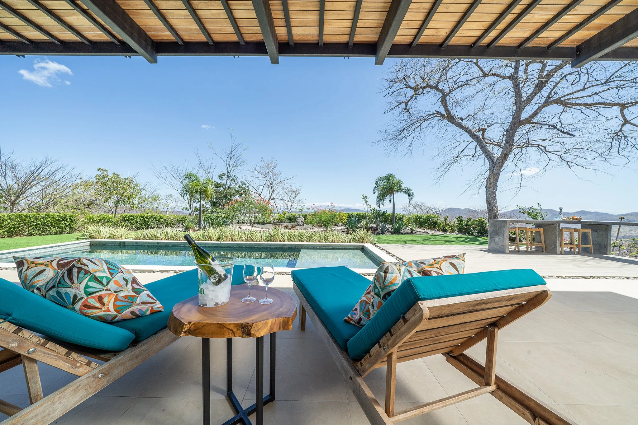 Outdoor patio with turquoise cushions and colorful pillows around a wooden table with a bottle of wine and two glasses, overlooking a yard with trees and mountains under a blue sky with clouds.