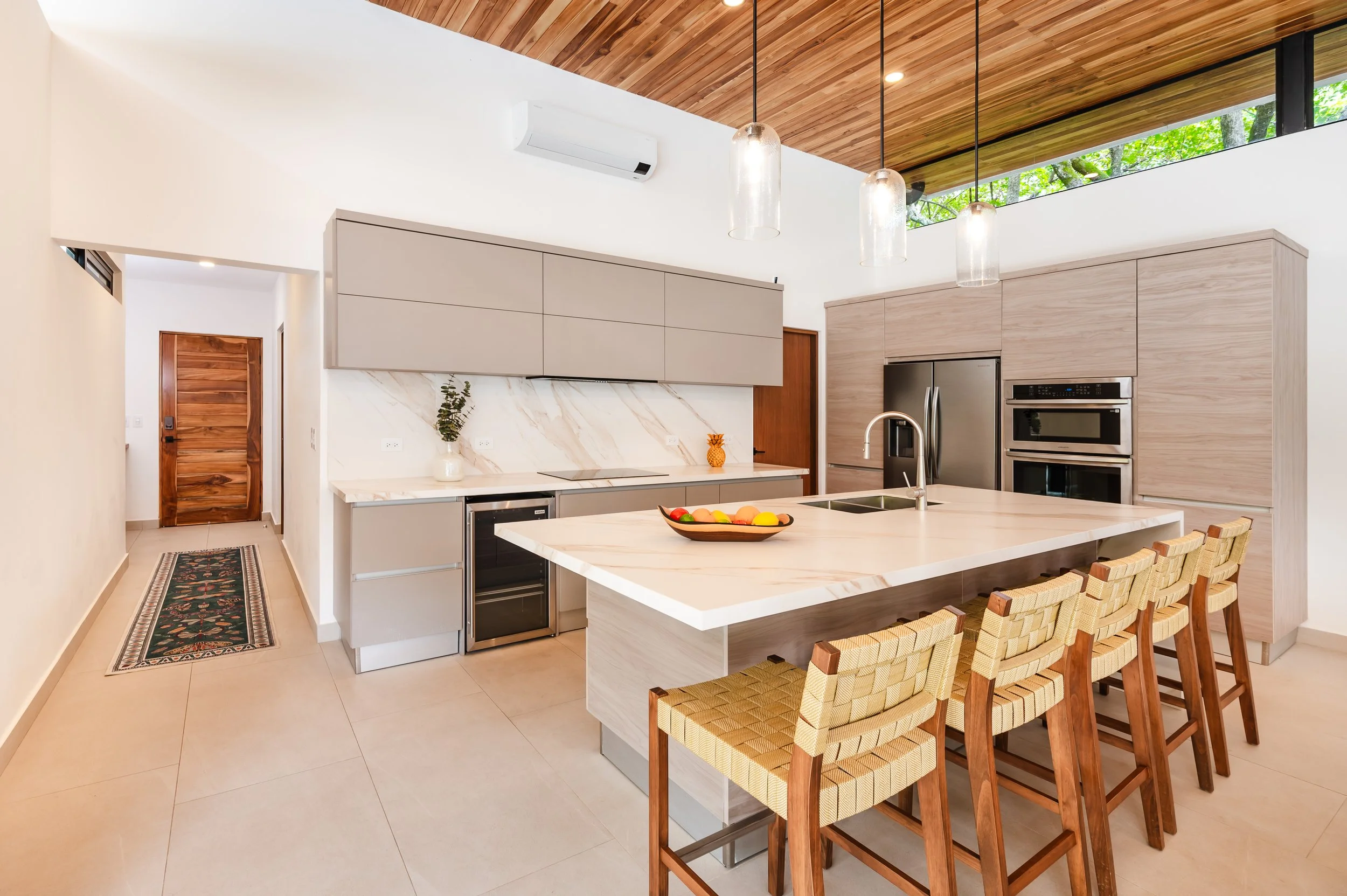Modern kitchen with beige cabinets, marble backsplash, and a kitchen island with woven chairs. Pendant lights hang from a wooden ceiling with large windows showing green trees.