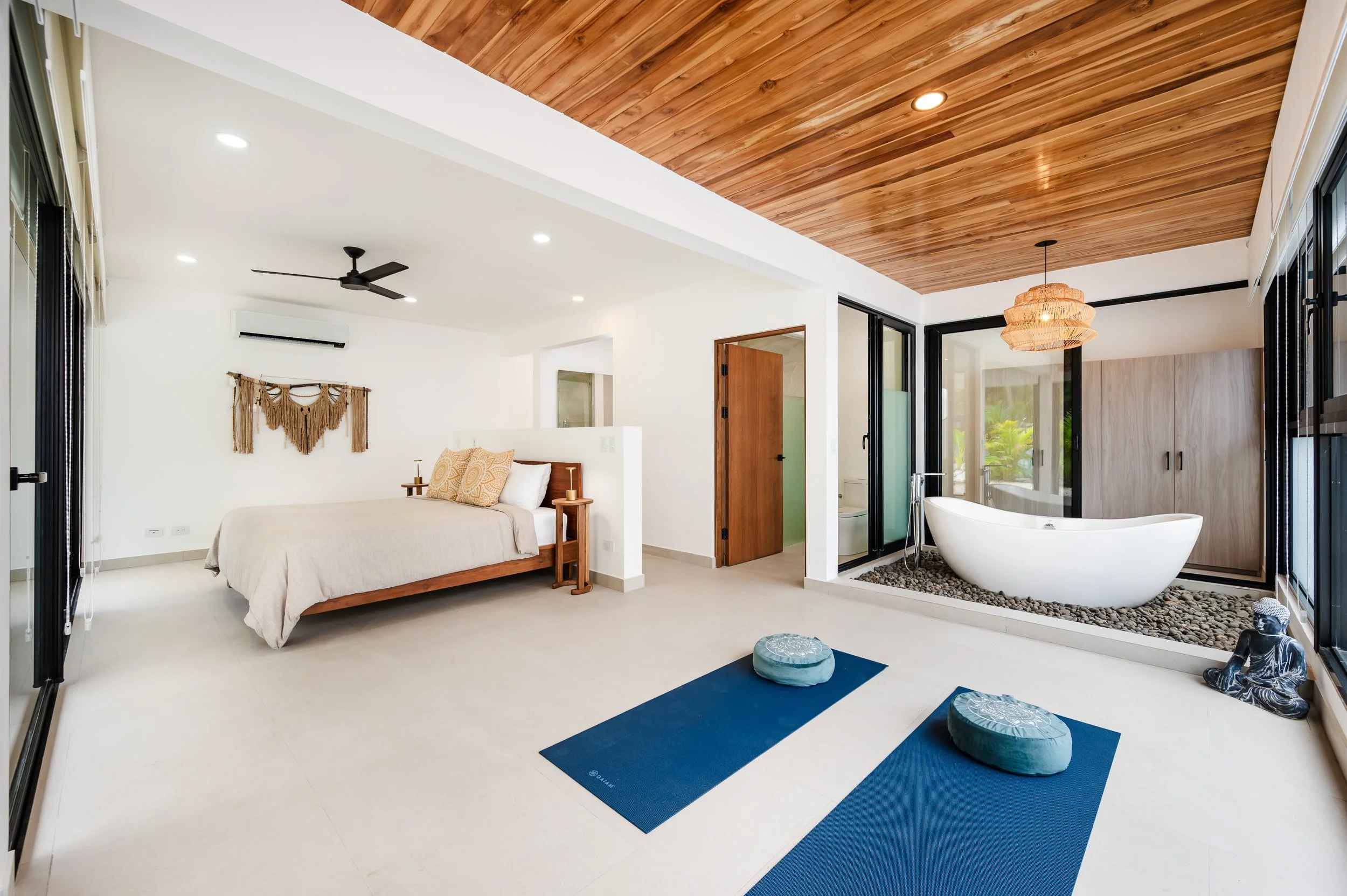 A bedroom with a wooden ceiling, a bed with beige bedding, a macrame wall hanging, a black ceiling fan, and white walls. There are large sliding glass doors leading to a bathroom with a white bathtub and a separate shower with glass doors, surrounded