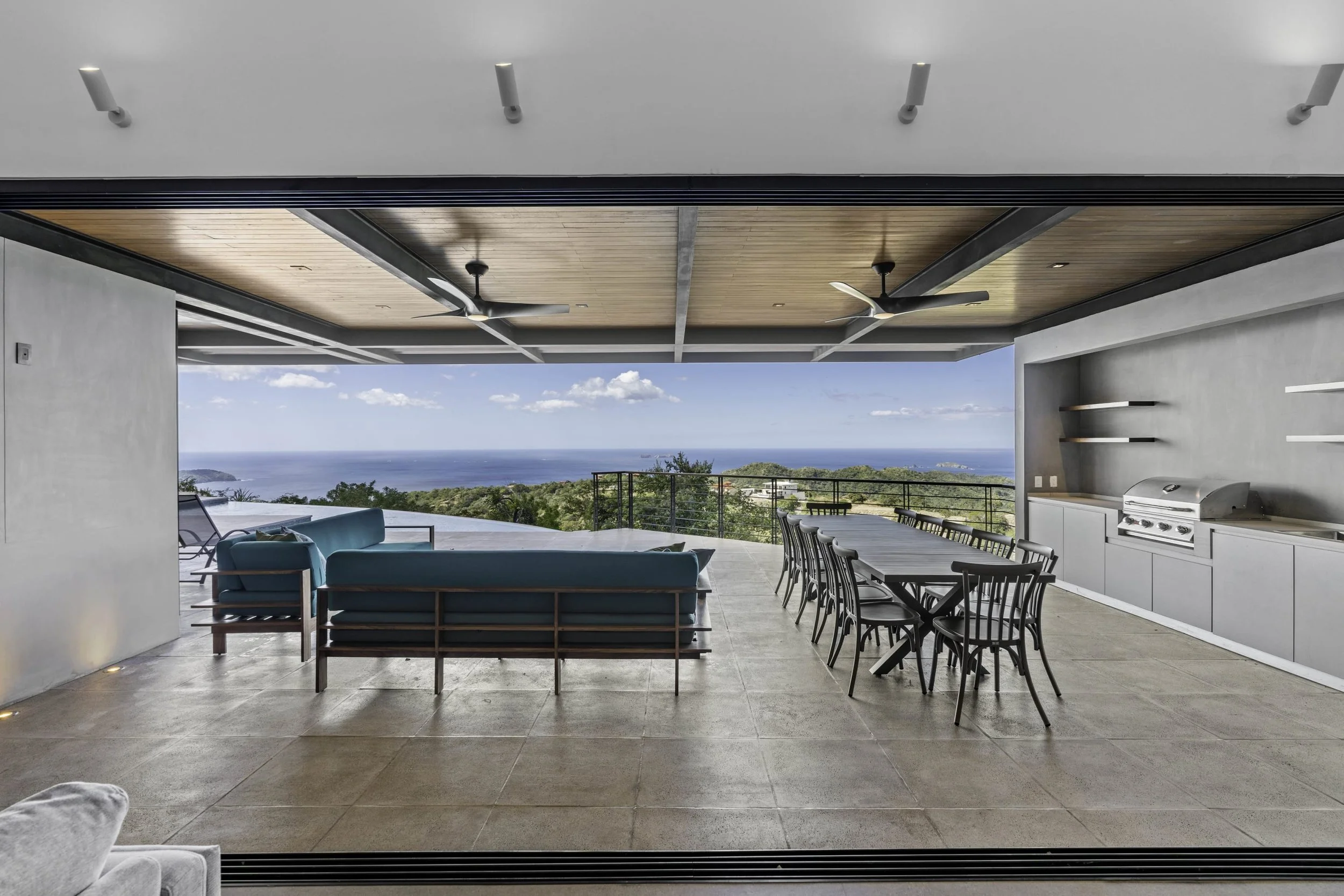 Modern outdoor living area with a view of the ocean, featuring a wooden ceiling with ceiling fans, a dining table with chairs, a grill, and a seating area with turquoise sofas next to a pool.