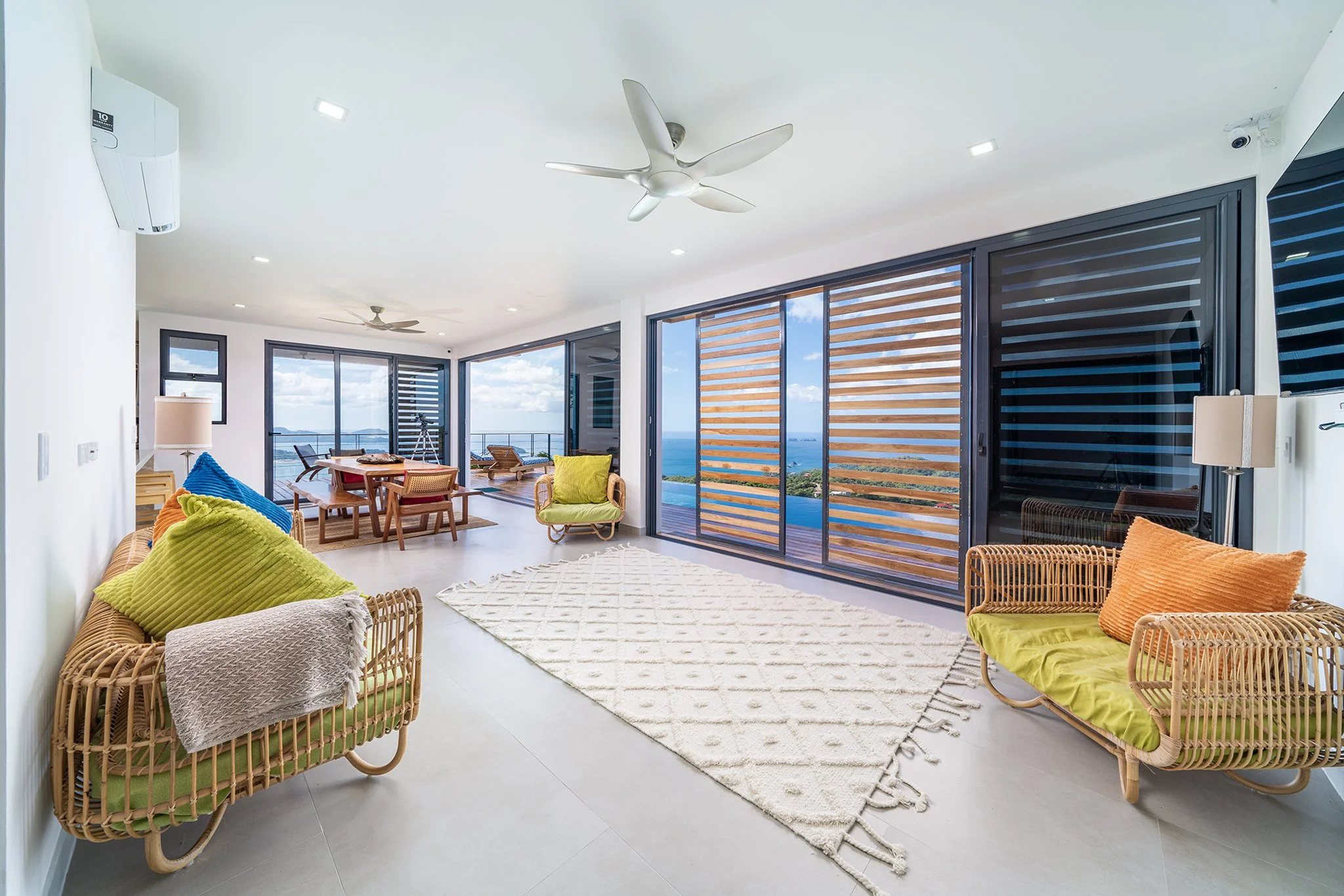 Bright and airy living room with large glass sliding doors that reveal a scenic view of the ocean, featuring wicker furniture with colorful cushions, a white rug, and modern ceiling fans.