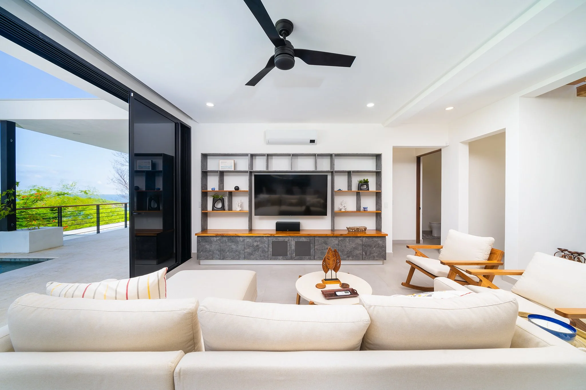 Living room with white sofas, a wooden coffee table, built-in media unit with a TV, and open sliding glass doors leading to an outdoor balcony with green foliage and trees.