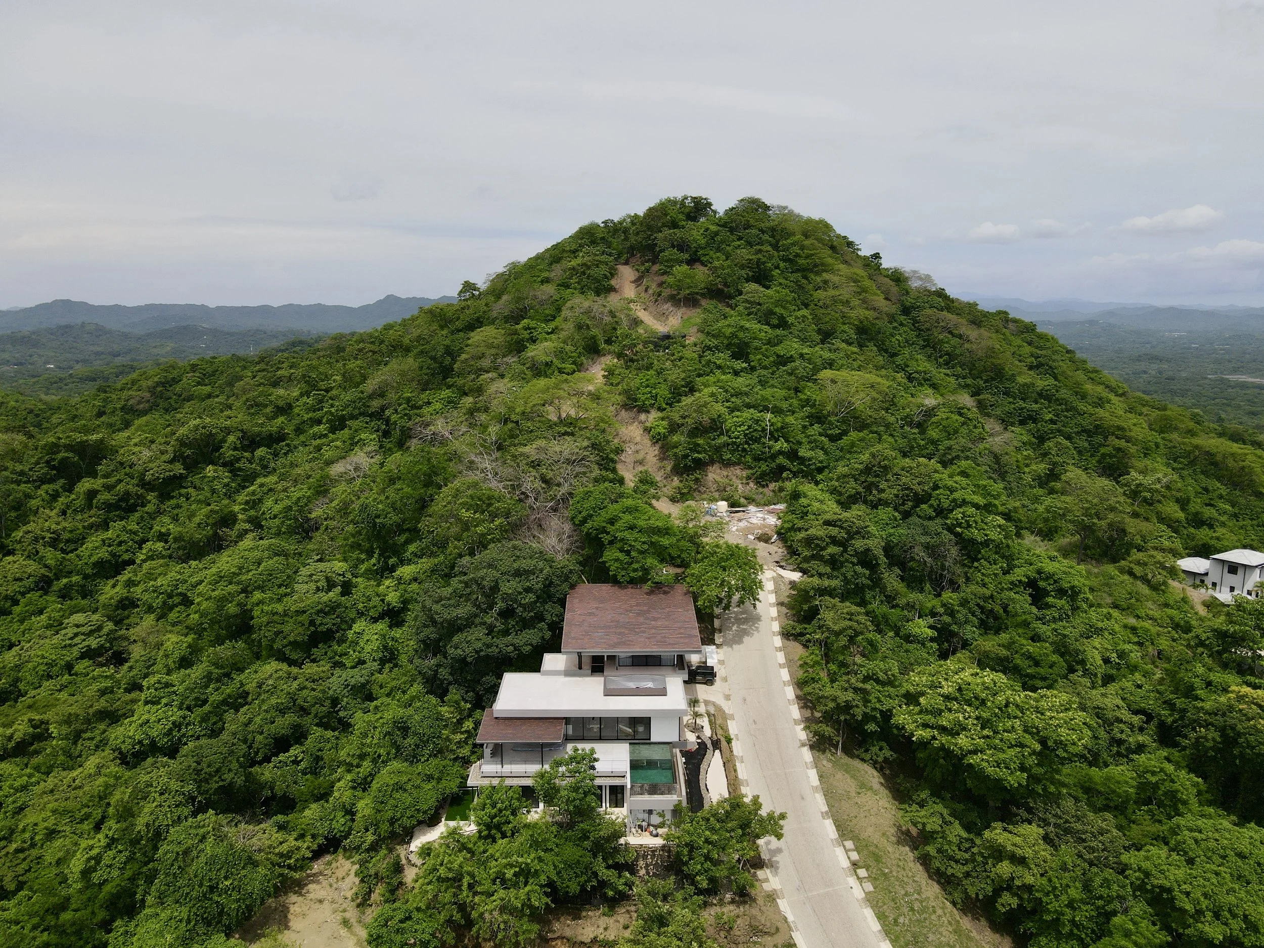 Aerial view of a modern house built on a lush green hillside, with a driveway leading to the entrance, surrounded by dense trees and forested mountains in the background.
