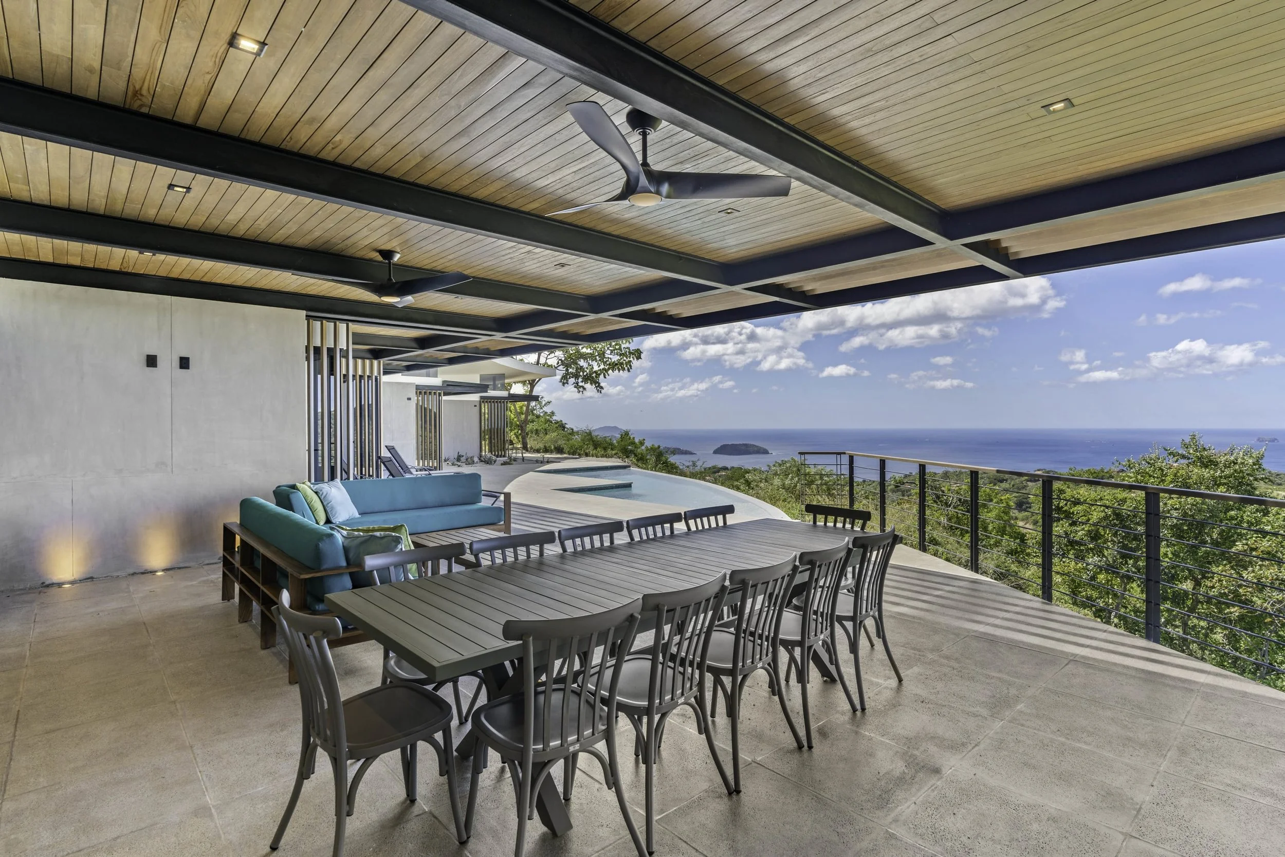 A modern outdoor patio with a wooden ceiling and ceiling fans, featuring a lounge area with a turquoise sofa, a dining table with several chairs, and a view of the ocean and islands in the distance.