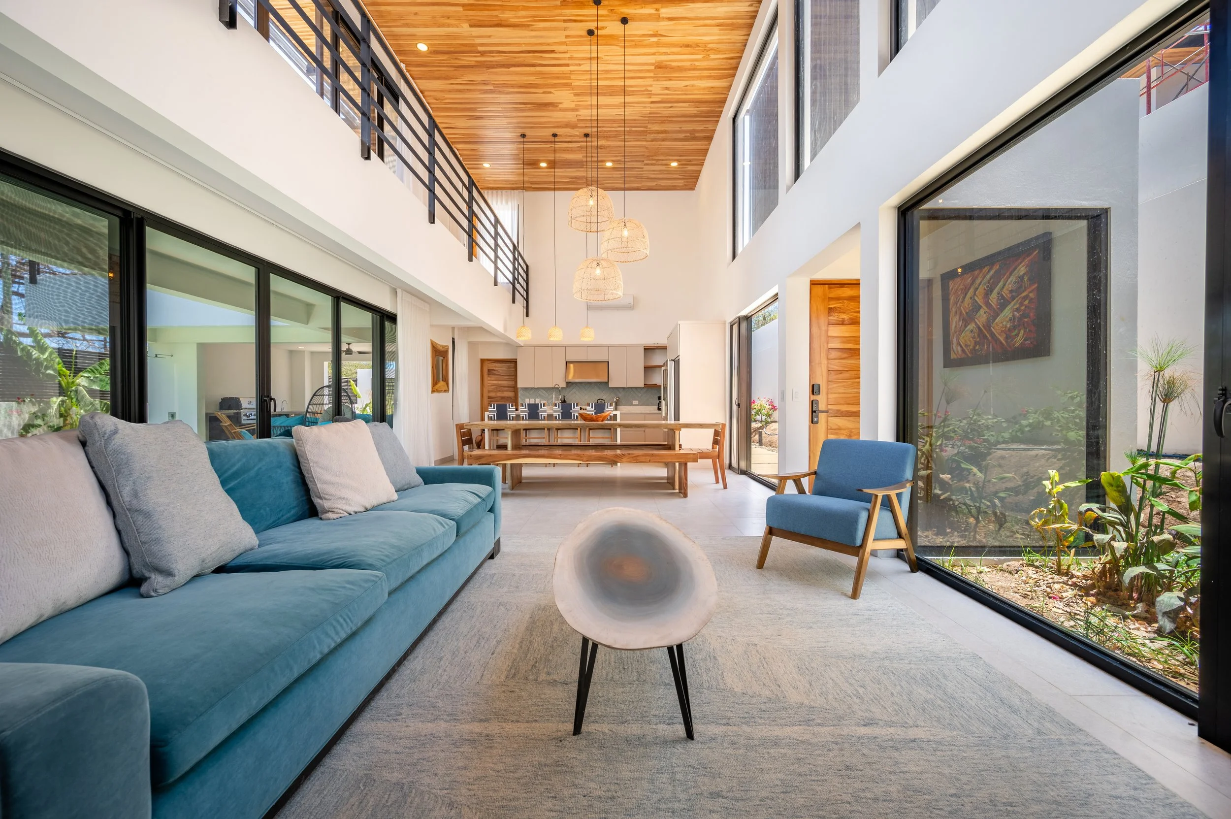 Interior view of a modern living room with large floor-to-ceiling windows, a blue sofa with multiple pillows, a light-colored rug, and a round wooden coffee table. The space opens up to a dining and kitchen area with wooden accents, pendant lights, a