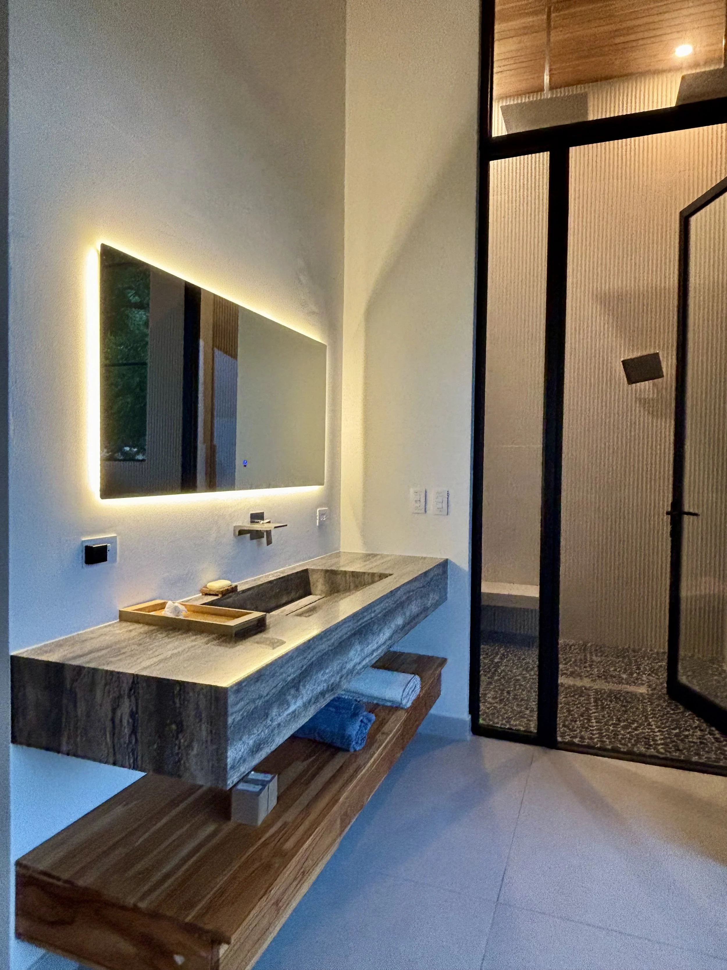 Modern bathroom with illuminated mirror, stone sink, wooden shelf, and textured glass door.