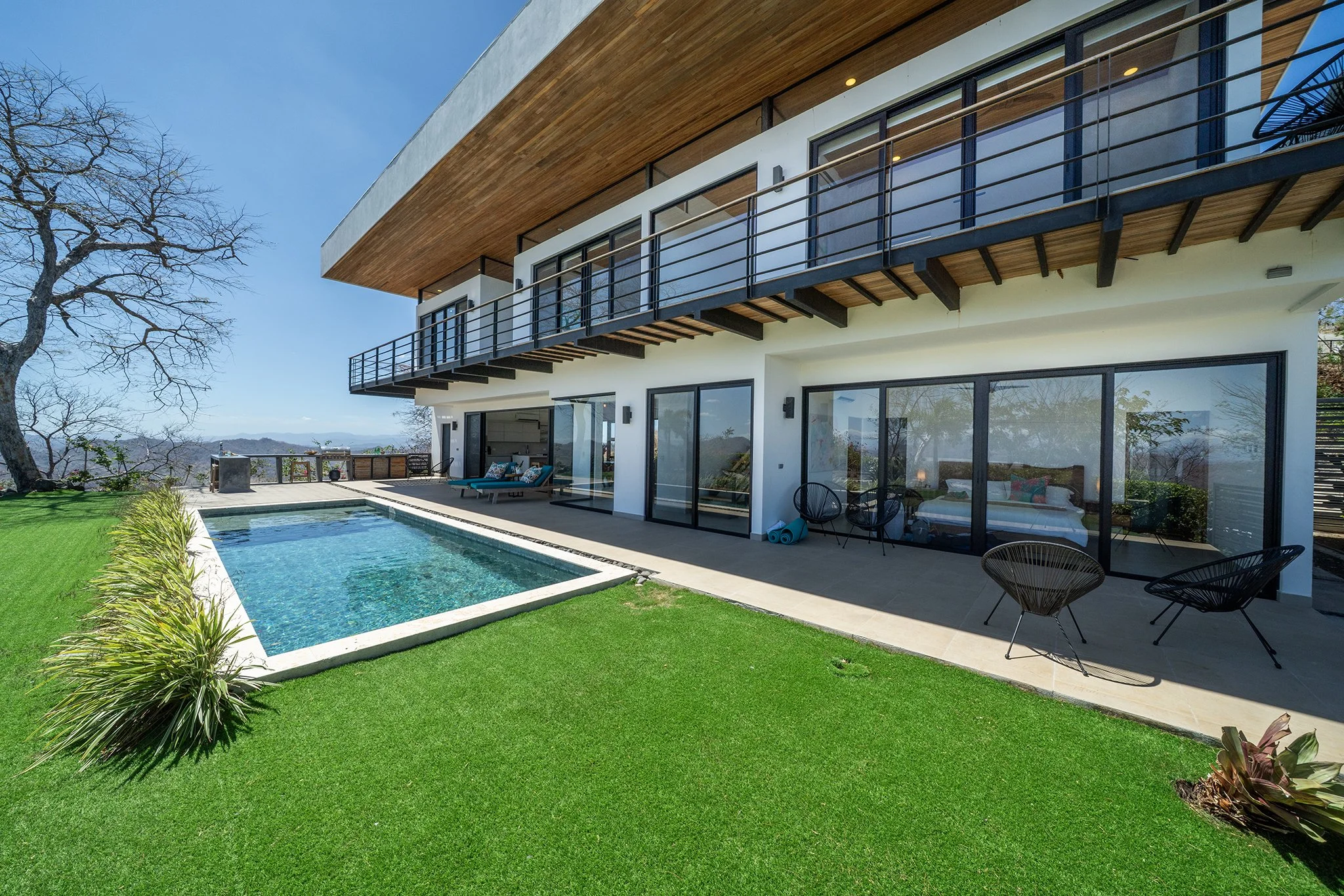 Modern house with large glass sliding doors, a swimming pool, and a patio with outdoor furniture, set in a lush green yard with a tree on the left side and a clear blue sky.