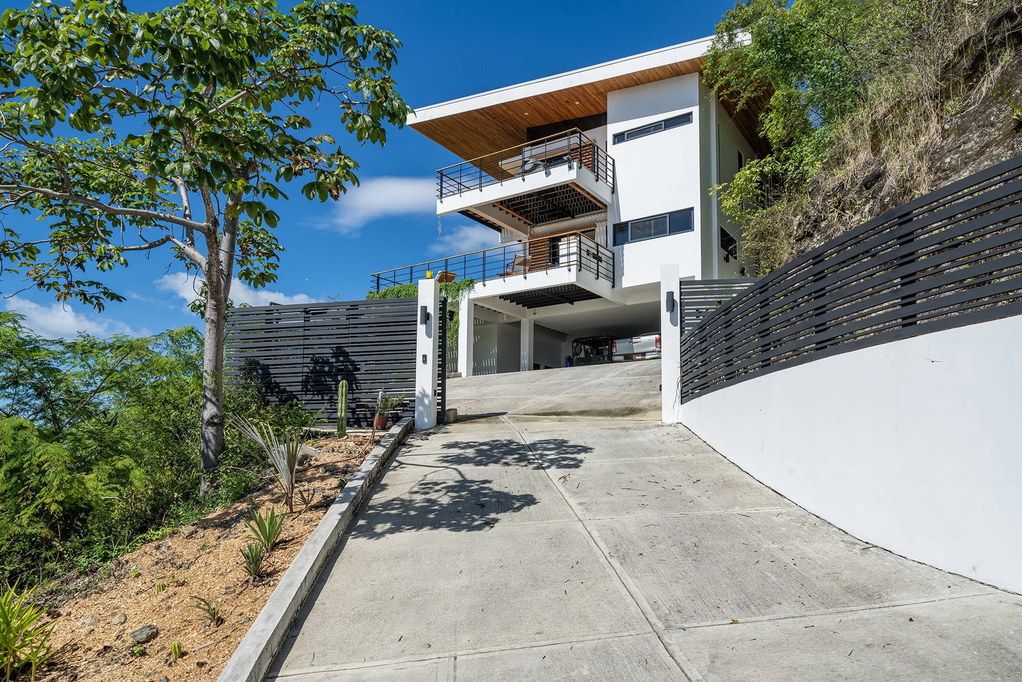 Modern white house with multiple balconies, black railings, and a sloped roof, situated on a hill with a concrete driveway and a lush green landscape under a blue sky.