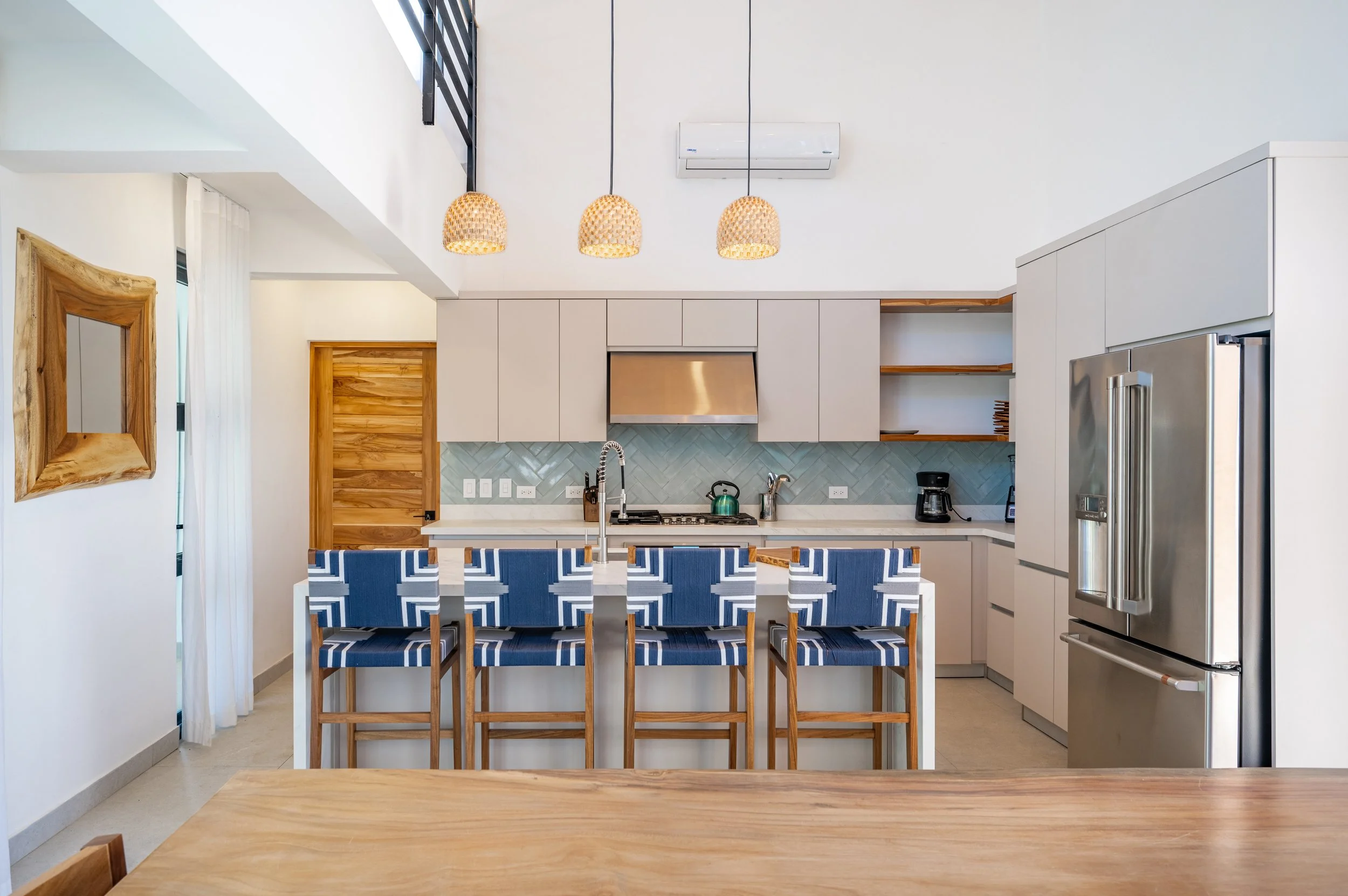 Modern kitchen with white cabinets, stainless steel refrigerator, light wood accents, pendant lights, and a kitchen island with blue chairs.