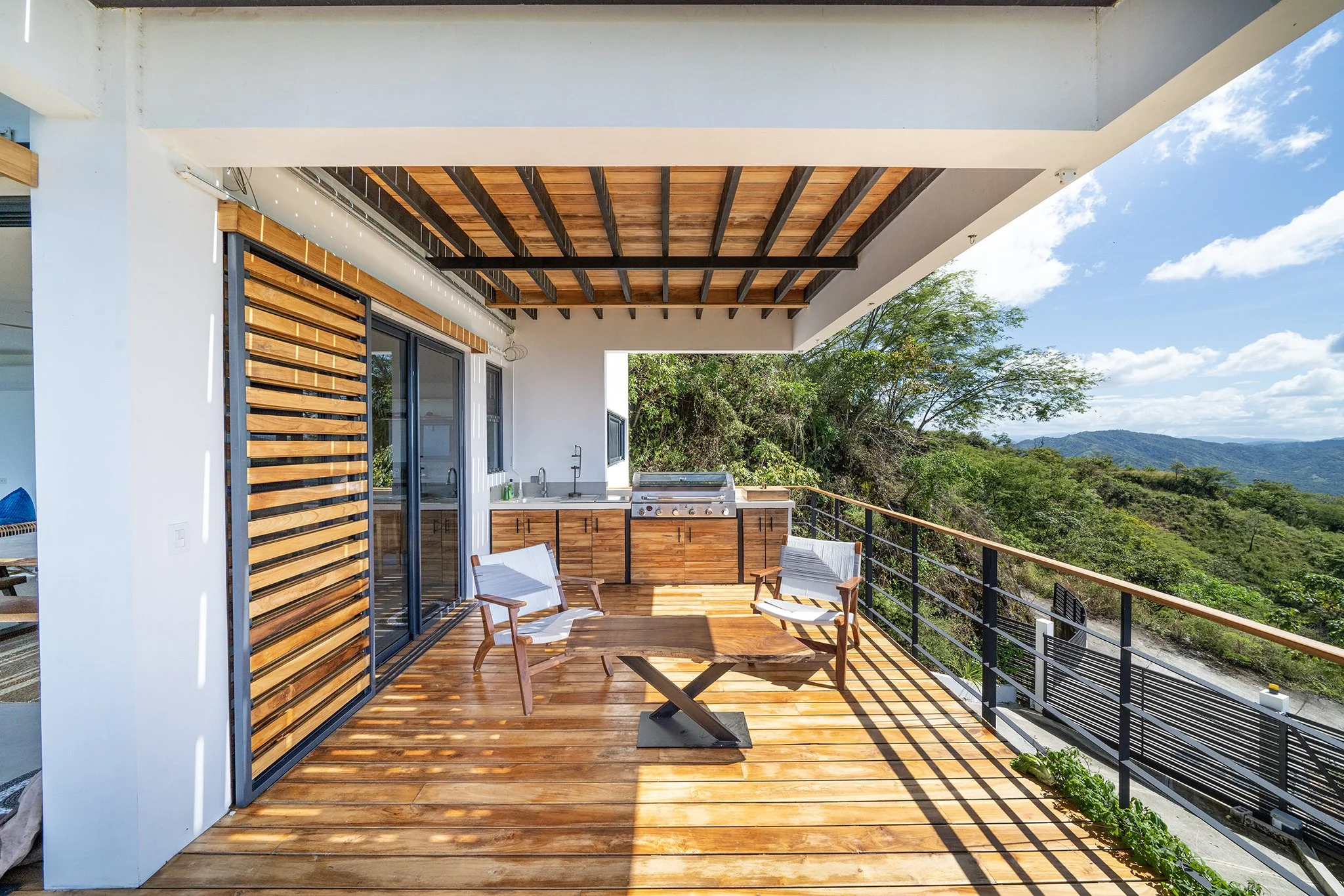 Modern outdoor balcony with wooden flooring and furniture, metal railing, built-in kitchen area with sink and grill, view of green trees and mountains under blue sky with clouds.