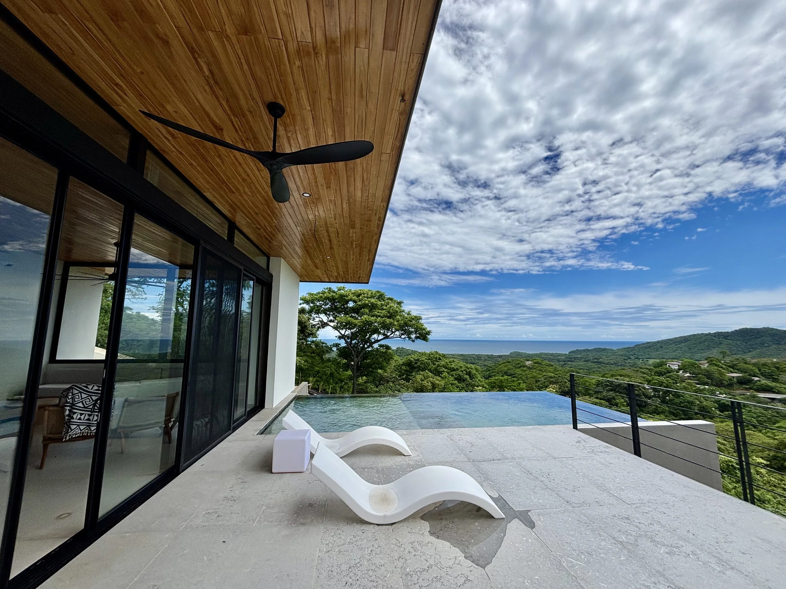 Patio area with white lounge chairs, overlooking a scenic landscape with green trees, hills, and a cloudy sky, adjacent to a modern house with large glass windows and wooden ceiling.