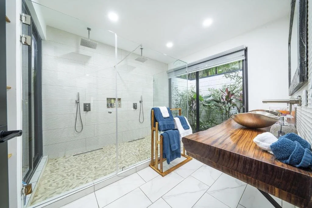 Modern bathroom with a glass-enclosed shower featuring two shower heads, a wooden countertop with a metallic vessel sink, and towels. Large windows show greenery outside.