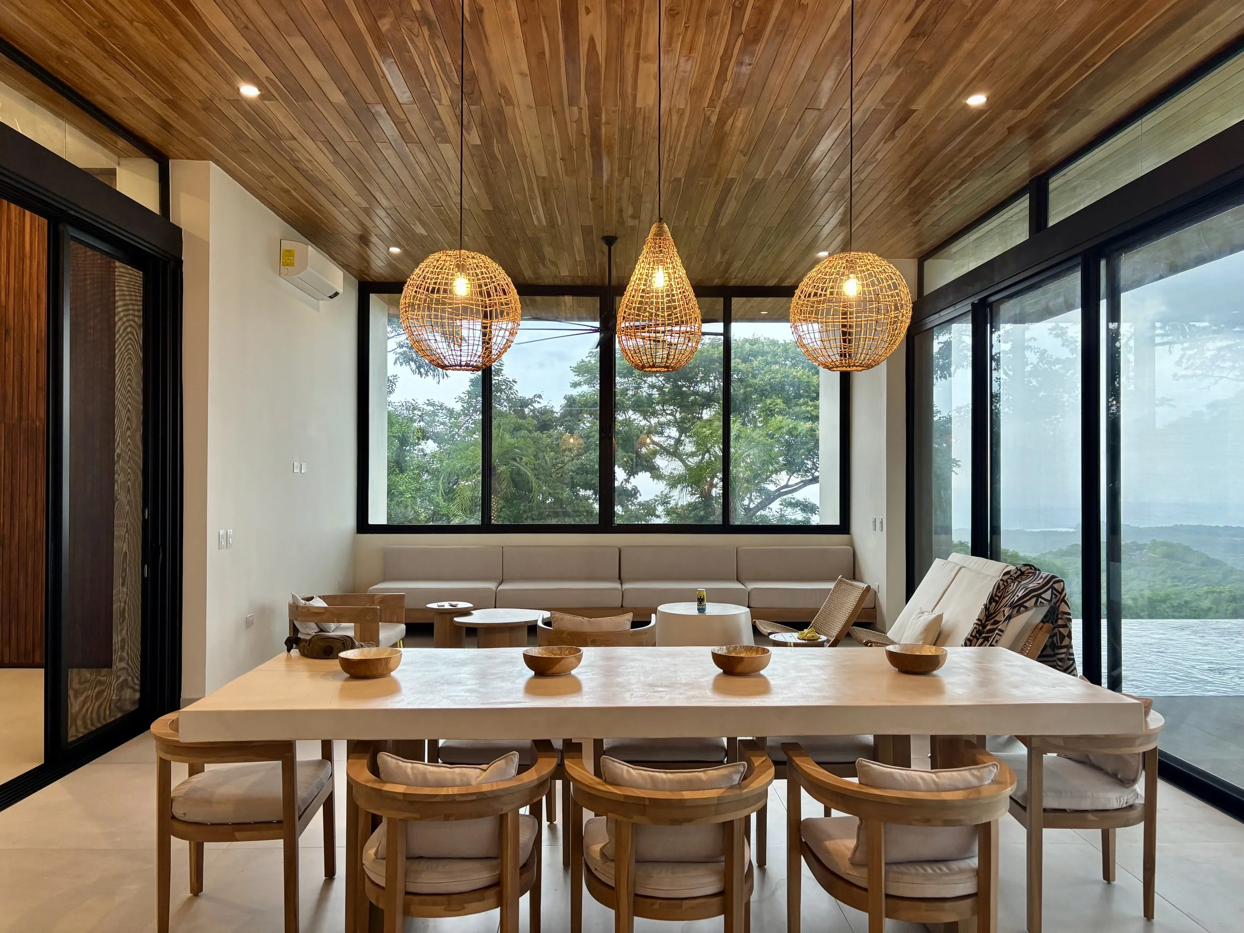 Modern dining area with large wooden table, surrounded by chairs, with a view of greenery through floor-to-ceiling windows, and decorated with three wicker pendant lights hanging from a wooden ceiling.
