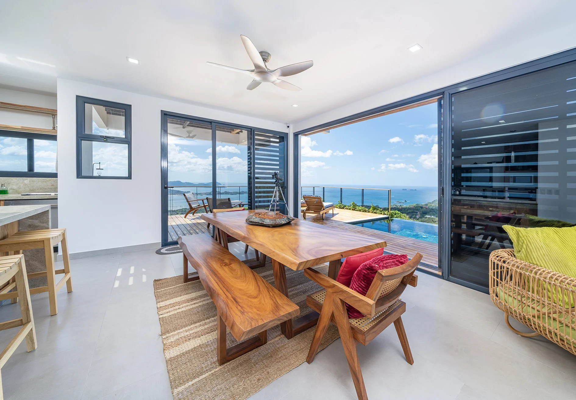 Modern living area with large glass sliding doors opening to a balcony overlooking the ocean. Wooden dining table with bench and chairs, ceiling fan, and ocean view.