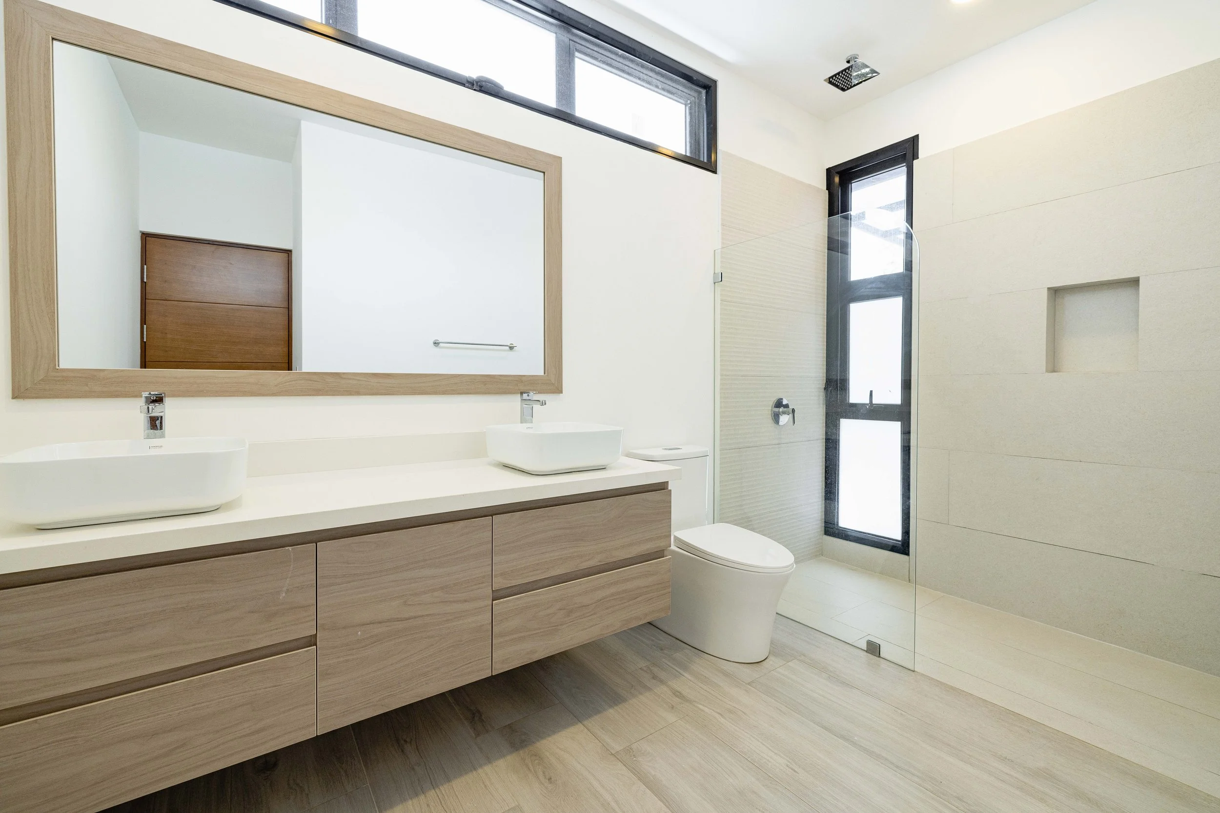 Modern bathroom with a large mirror, floating vanity with two vessel sinks, a toilet, a walk-in glass shower with a rainfall showerhead, and high windows for natural light.