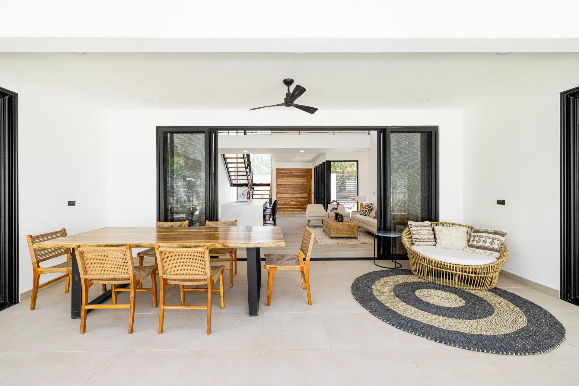Open-plan living and dining area with a large wooden dining table, rattan chairs, a rug, a beige sofa, armchair, and black sliding doors leading to a modern kitchen and staircase.