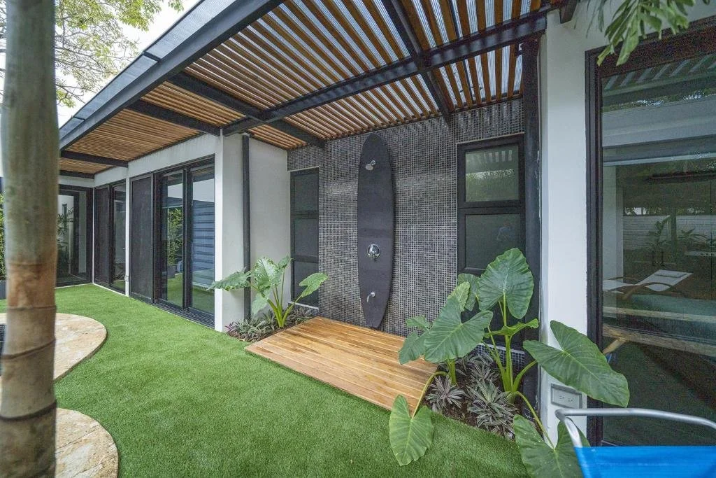 Outdoor patio area with a wooden deck and grass flooring, glass sliding doors, some leafy potted plants, and a black surfboard-shaped water feature attached to the wall.