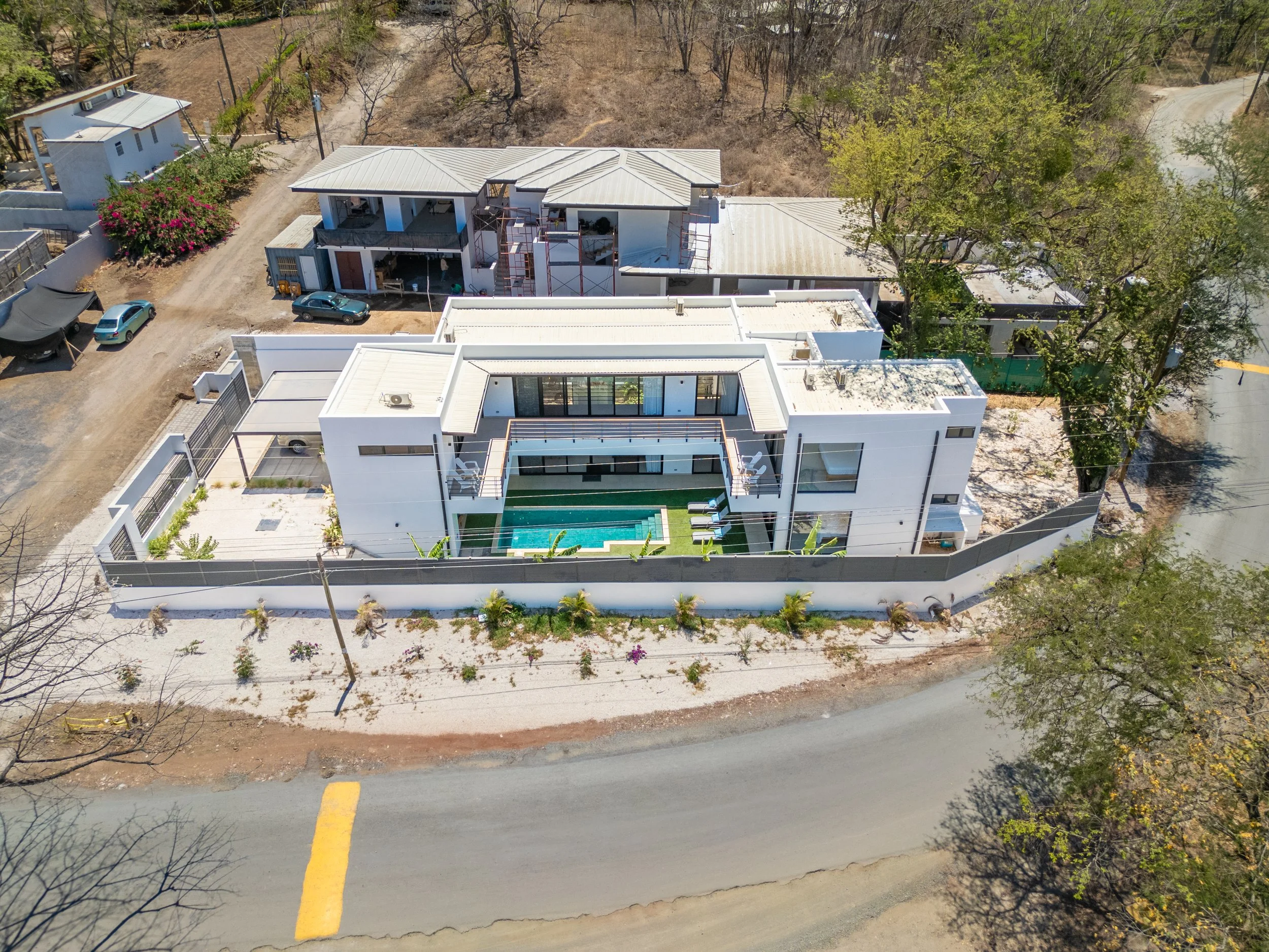 Aerial view of a modern white house under construction with scaffolding on sides, a swimming pool, and a backyard with small trees and shrubs, surrounded by a white wall and nearby roads.