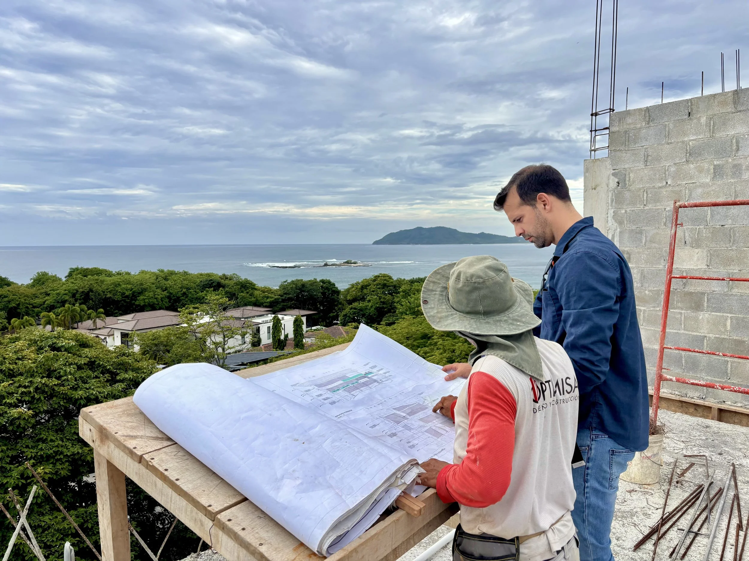 Two men reviewing blueprints on a construction site with a view of the ocean and a hill in the background.