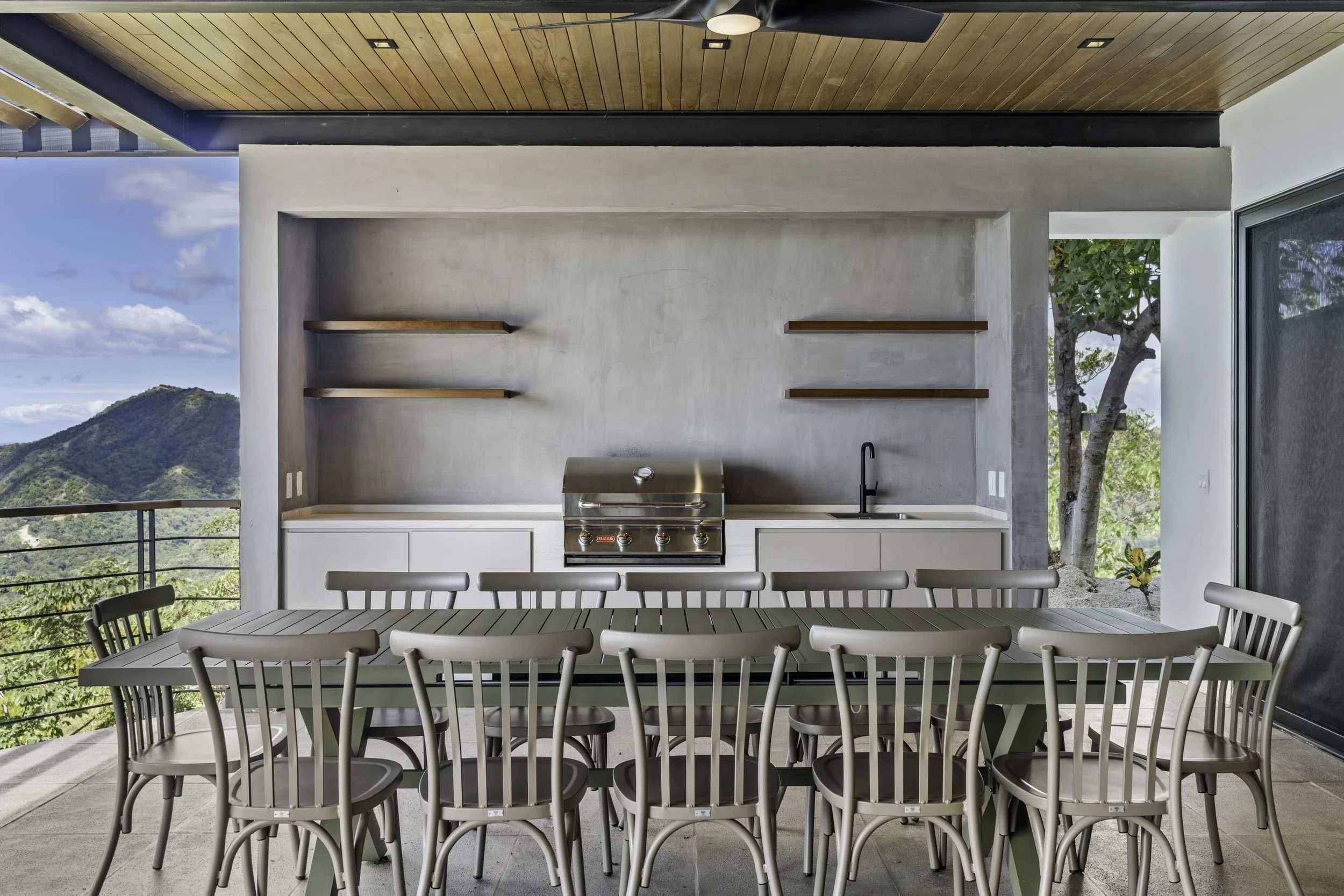 Outdoor dining area featuring a long table with chairs, a modern kitchen with a built-in grill, countertop, and sink, large sliding glass door, and scenic mountain view in the background under a wooden ceiling.