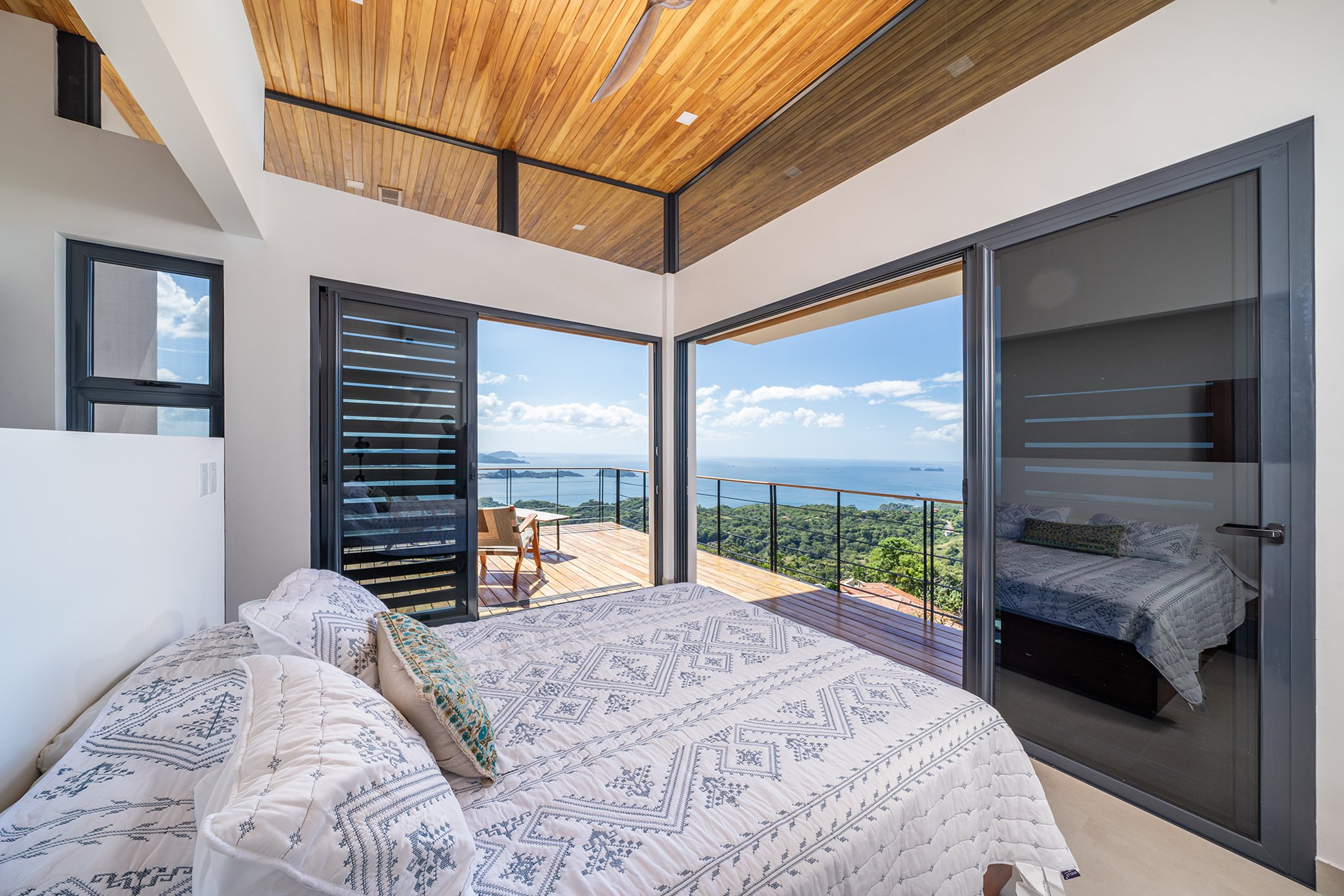 Bedroom with white bedding and decorative pillows, large sliding glass doors opening to a balcony with a view of the ocean and distant islands, wooden ceiling, modern design.