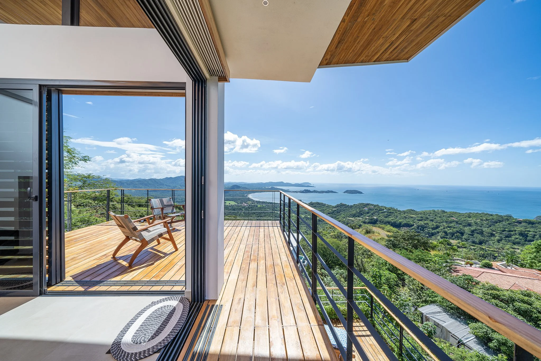 View from a modern balcony with wooden flooring, overlooking green hills and the ocean, with a clear blue sky and some clouds.