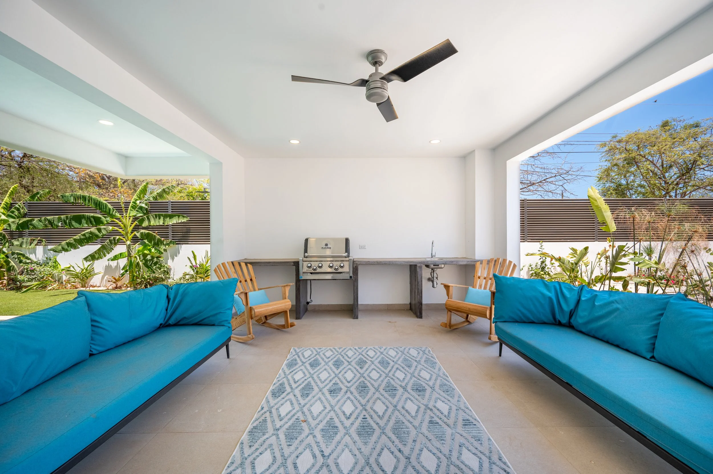 Outdoor covered patio with blue cushioned sofas, wooden chairs, a patterned rug, a built-in barbecue, and lush green plants, with trees and a fence in the background.