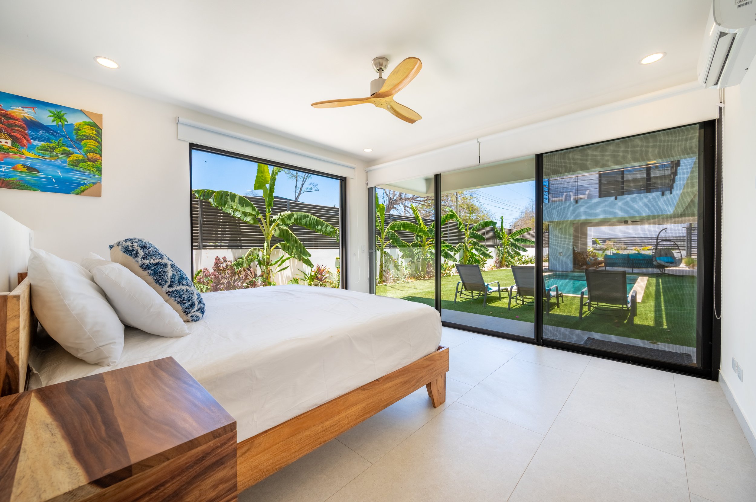 A bedroom with large glass sliding doors leading to a backyard with a pool and outdoor seating. The room features a wooden bed with white bedding and a blue patterned pillow, a wooden side table, a ceiling fan, and a colorful painting on the wall.